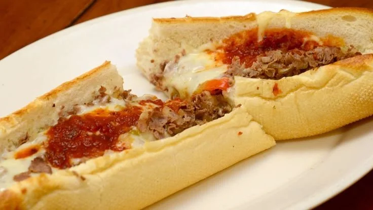 Philly cheesesteak sandwich with beef, melted cheese, and tomato sauce on a bun.
