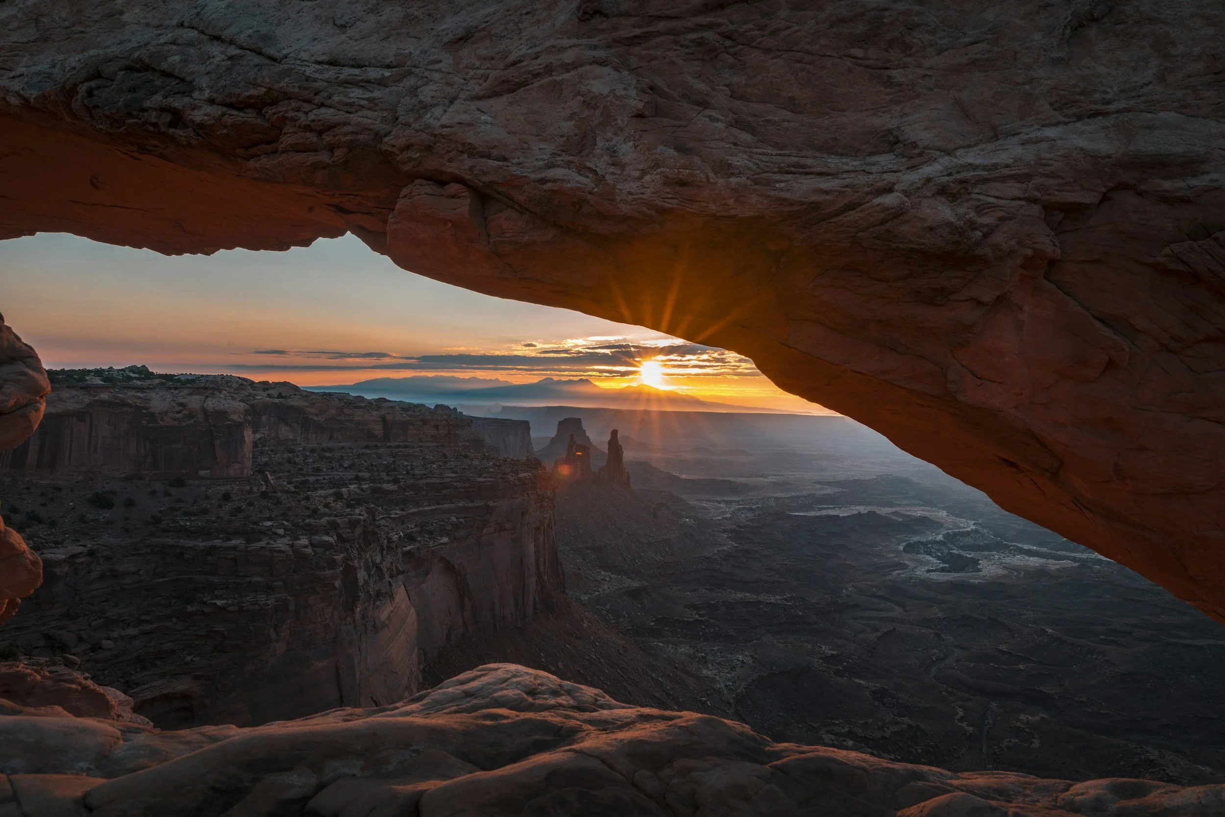 Sunset over a canyon viewed through a natural rock arch with rock formations in the distance.