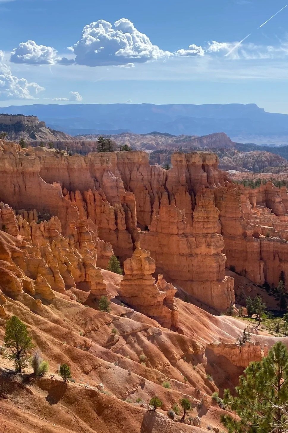 A panoramic view of the Bryce Canyon landscape featuring towering orange and pink hoodoos, sparse green trees, and distant layered cliffs under a bright blue sky with scattered clouds and a faint airplane contrail.