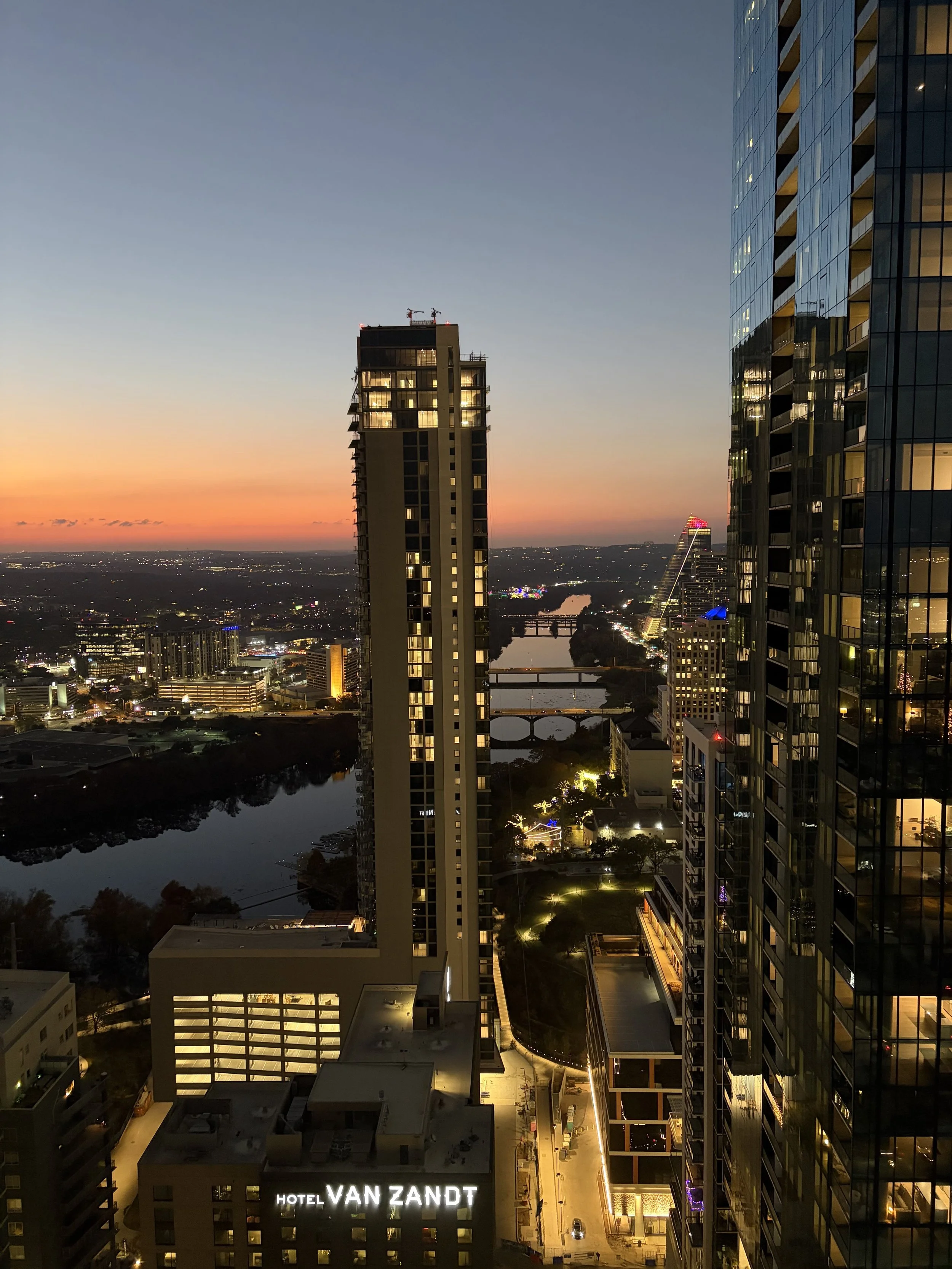 Cityscape at dusk with tall buildings, illuminated windows, a river, and a bridge, with a sunset sky in the background.