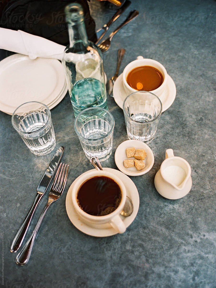 A table setting with two cups of black coffee, three glasses of water, a glass bottle of water, a small jug of cream, a small plate of sugar cubes, and fork and knife placed on a gray table surface.