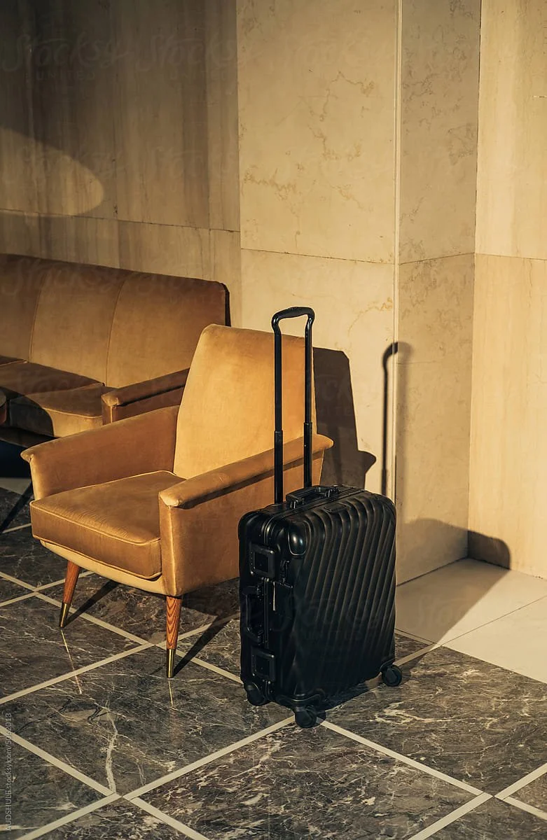 A black rolling suitcase with an extended handle next to an orange mid-century modern armchair in an indoor space with marble tile flooring and beige marble walls.
