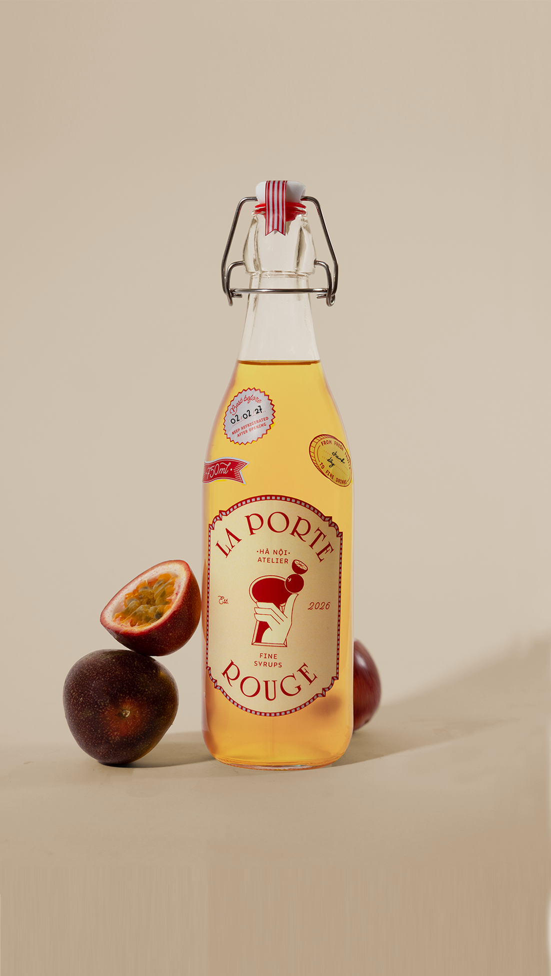 A glass bottle of syrup with a metal clasp, decorated with red and white striped ribbon, labeled 'La Porte Rouge' with a stylized hand holding a cherry. To the left, there is a halved passion fruit with yellow pulp and black seeds, and two whole passion fruits with dark purple skin.