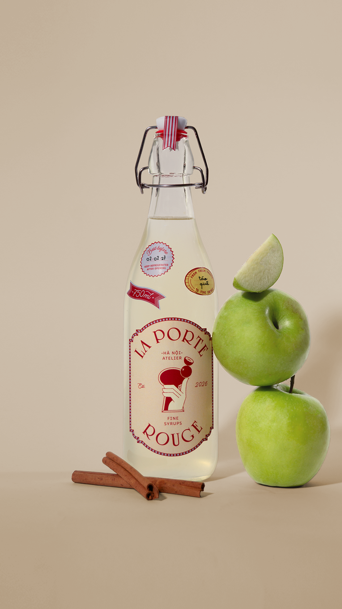 A clear glass bottle with a swing-top cap, labeled as La Porte Rouge, positioned next to three stacked green apples and two cinnamon sticks on a beige background.