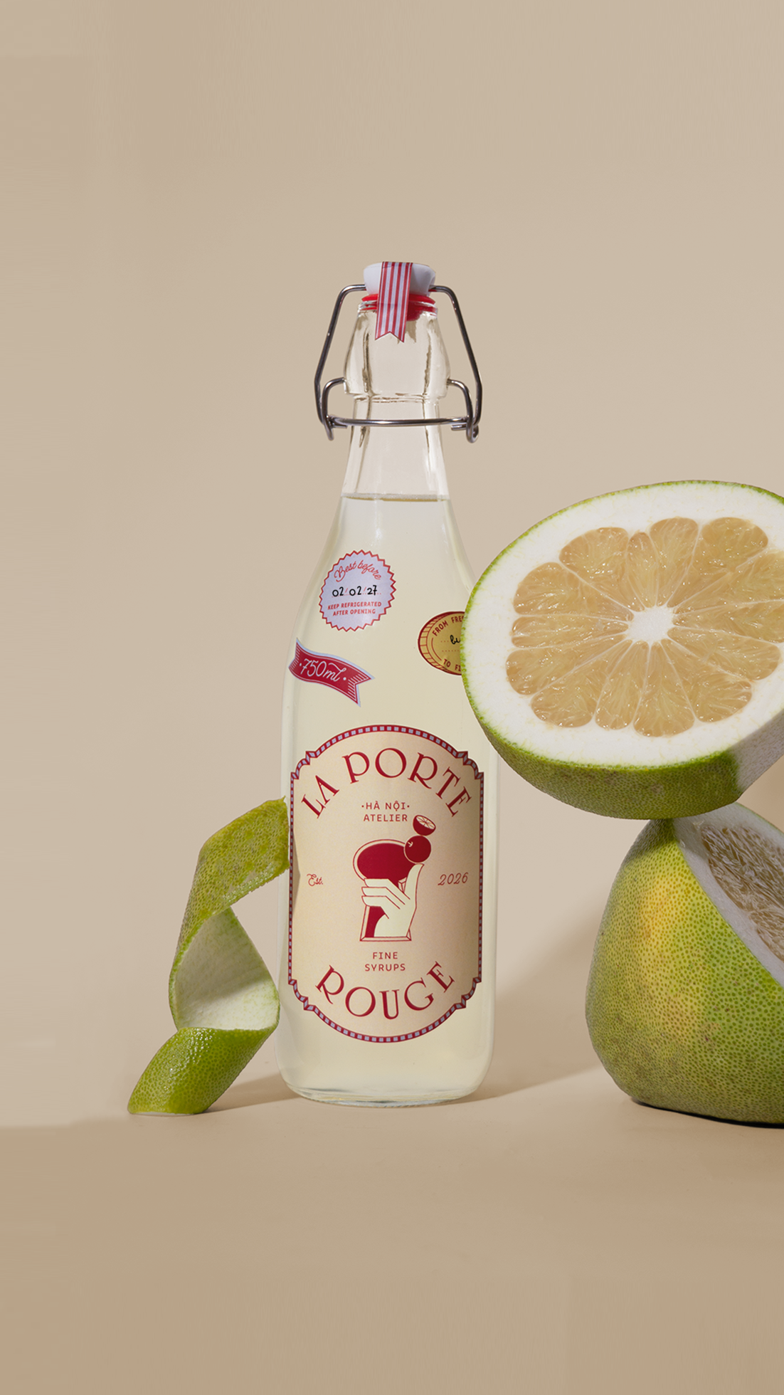 A glass bottle of lemon syrup with a red and white swing-top cap, surrounded by a halved lemon, a lemon peel peel, and a whole lemon, set against a beige background.