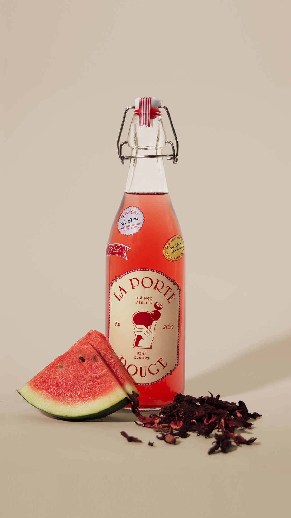 A glass bottle of pink syrup with a ceramic and metal clasp, labeled 'LA PORTE' with a watermelon illustration, placed beside a slice of watermelon and dried hibiscus petals on a light background.