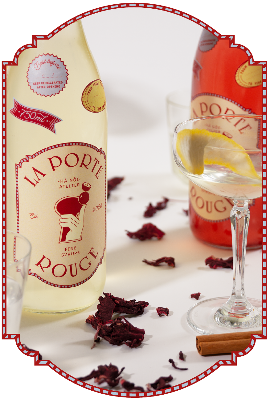 A bottle of La Porte Rouge syrup with a lemon wedge in a cocktail glass and a cinnamon stick on the table, surrounded by dried hibiscus petals.