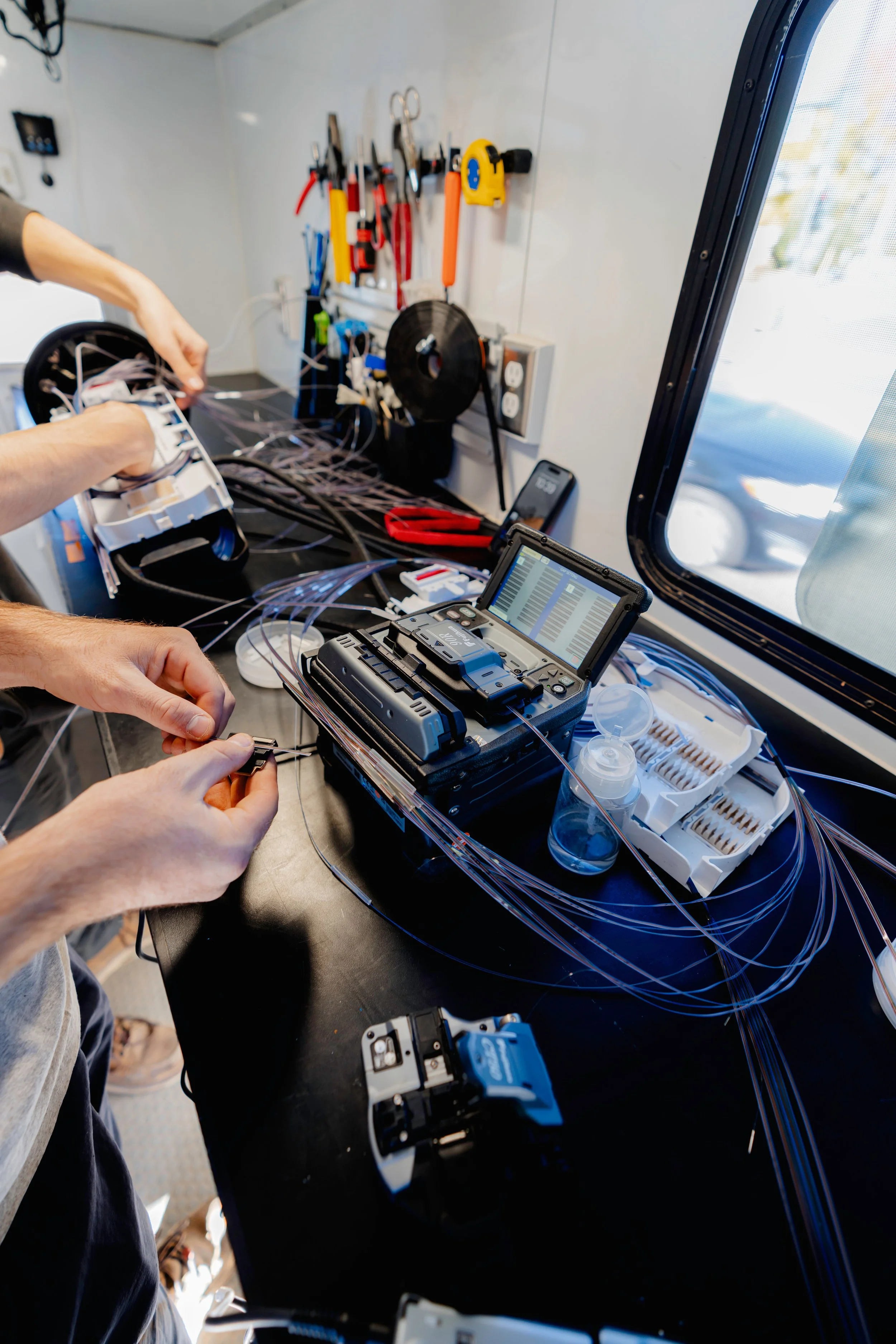 Individuals splicing fiber optic cable inside a fiber splicing trailer and showing tools on a black workstation.