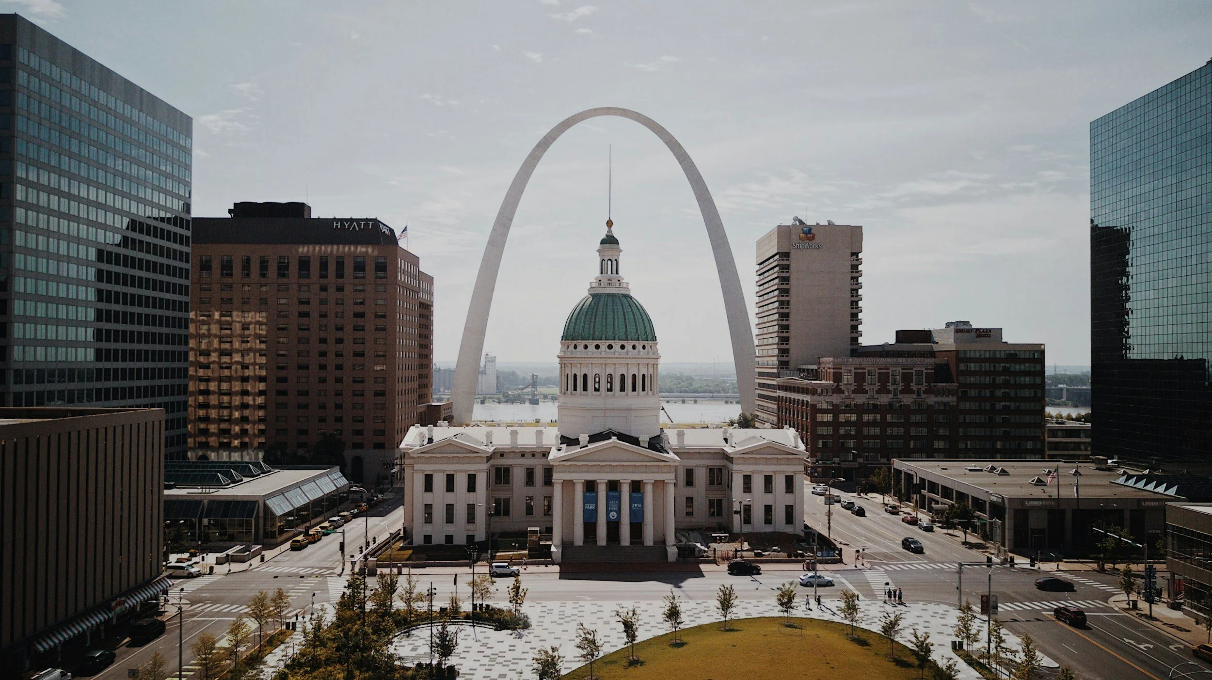 This image shows a cityscape with tall modern buildings and the Gateway Arch in the background, along with a historic building featuring a green dome and white columns in the foreground.