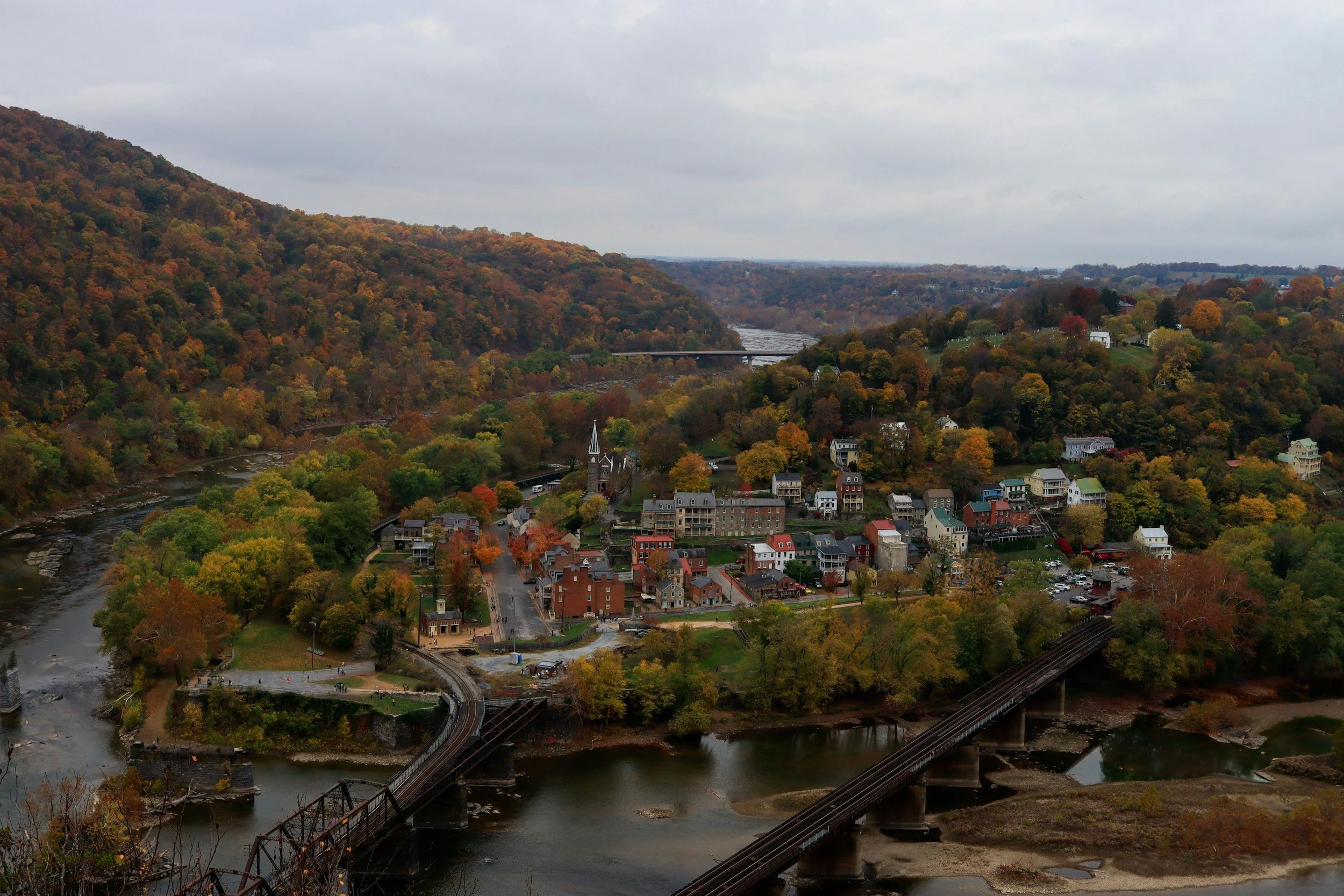 A scenic view of a town nestled in a valley surrounded by forested hills with fall foliage, a river flowing through, and bridges connecting different parts of the town under an overcast sky.