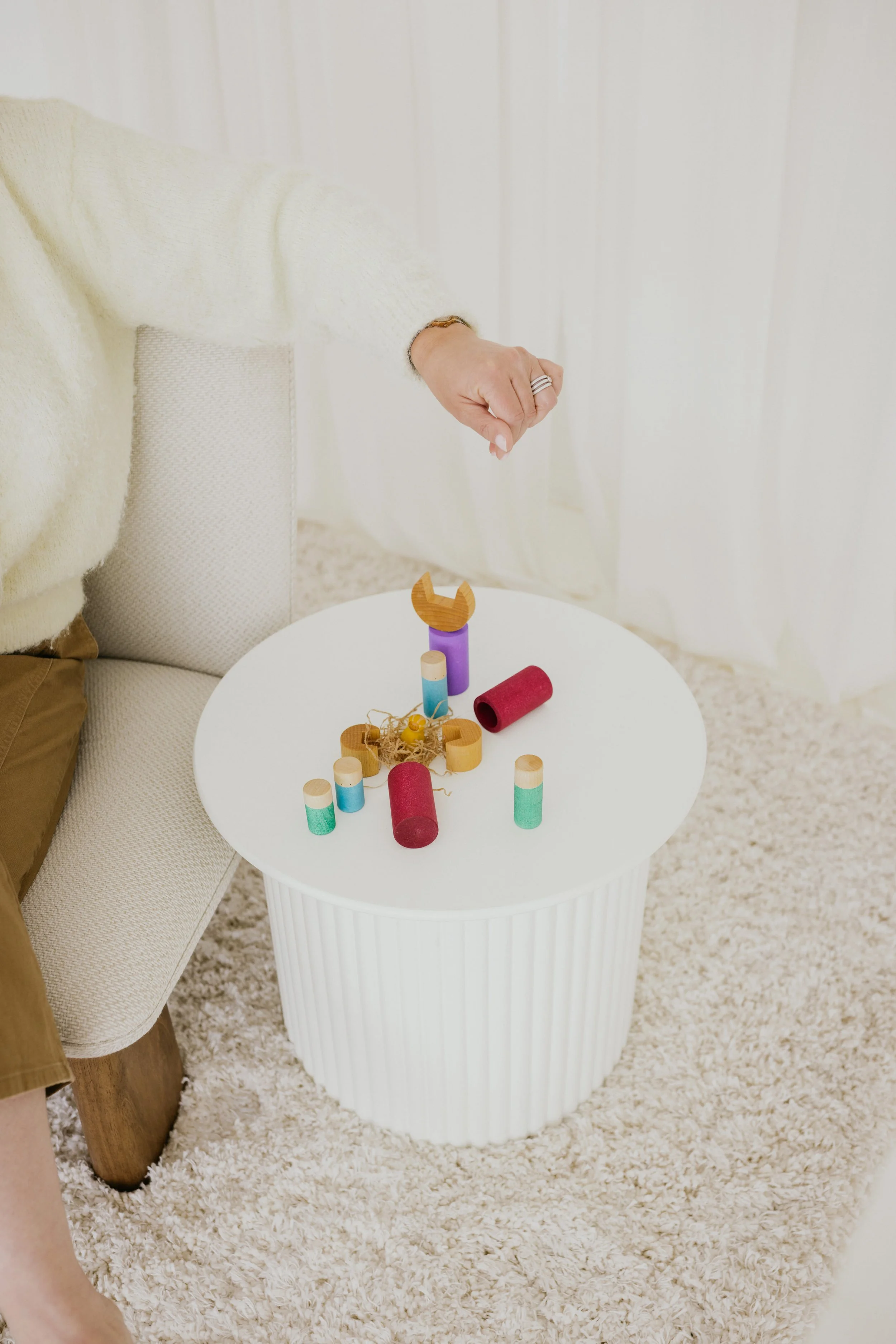 Een witte ronde tafel met houten en gekleurde blokken en een stuk touw, naast een beige bank en een iemand die een hand uitstrekt