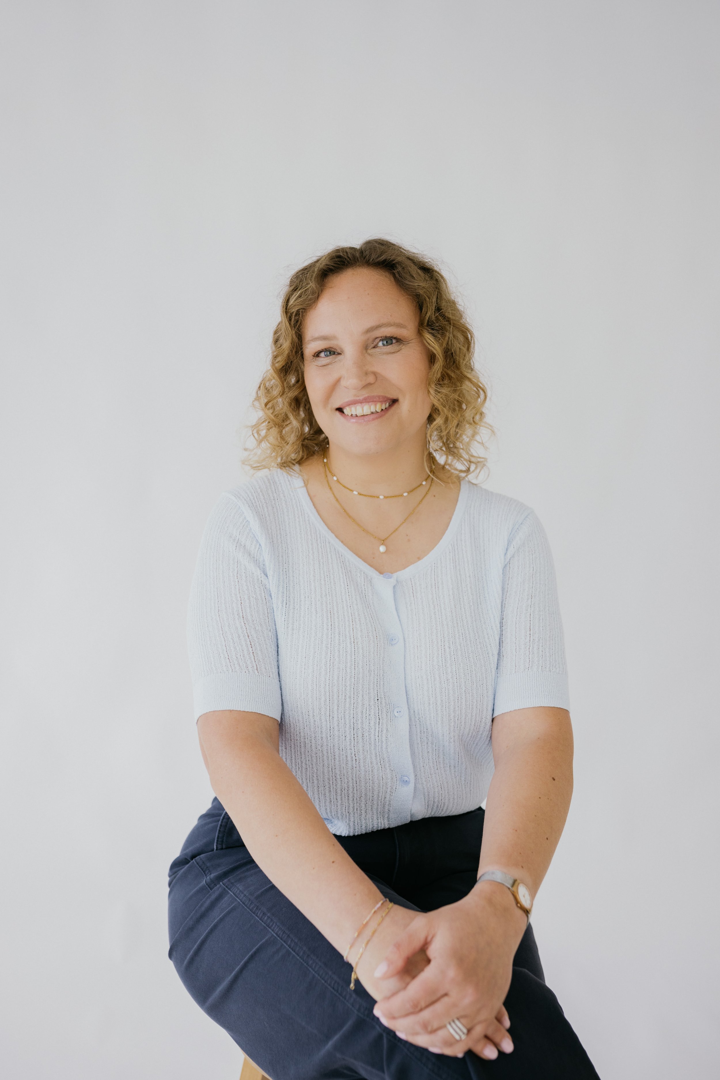 Een glimlachende vrouw met gekruld blond haar, gekleed in een lichtblauwe blouse en donkere broek, zittend met haar handen gevouwen op haar knie voor een witte achtergrond.