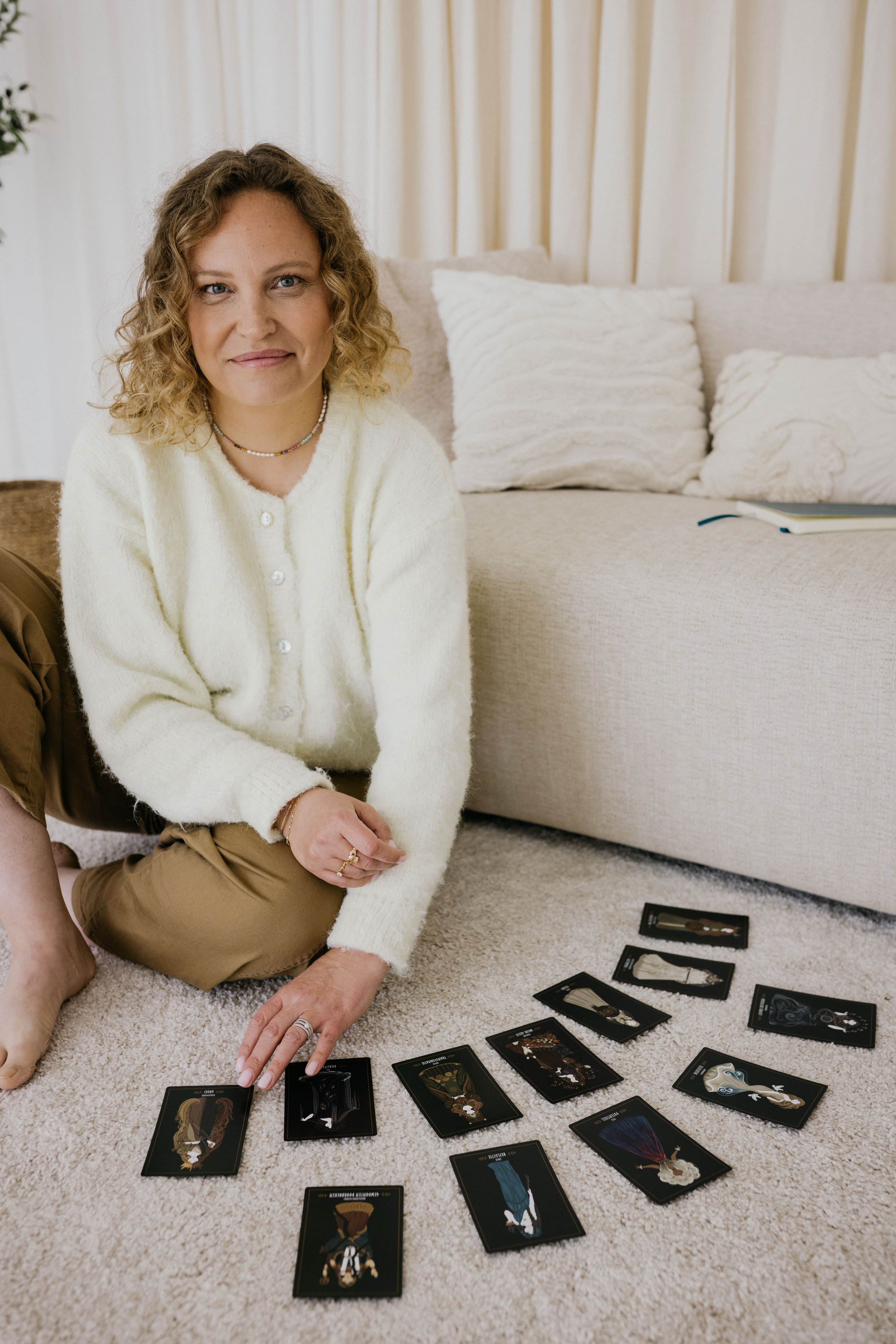 Vrouw zittend op tapijt met tarotkaarten uitgespreid voor haar, in een gezellige praktijkruimte met een bank en kussens op de achtergrond.