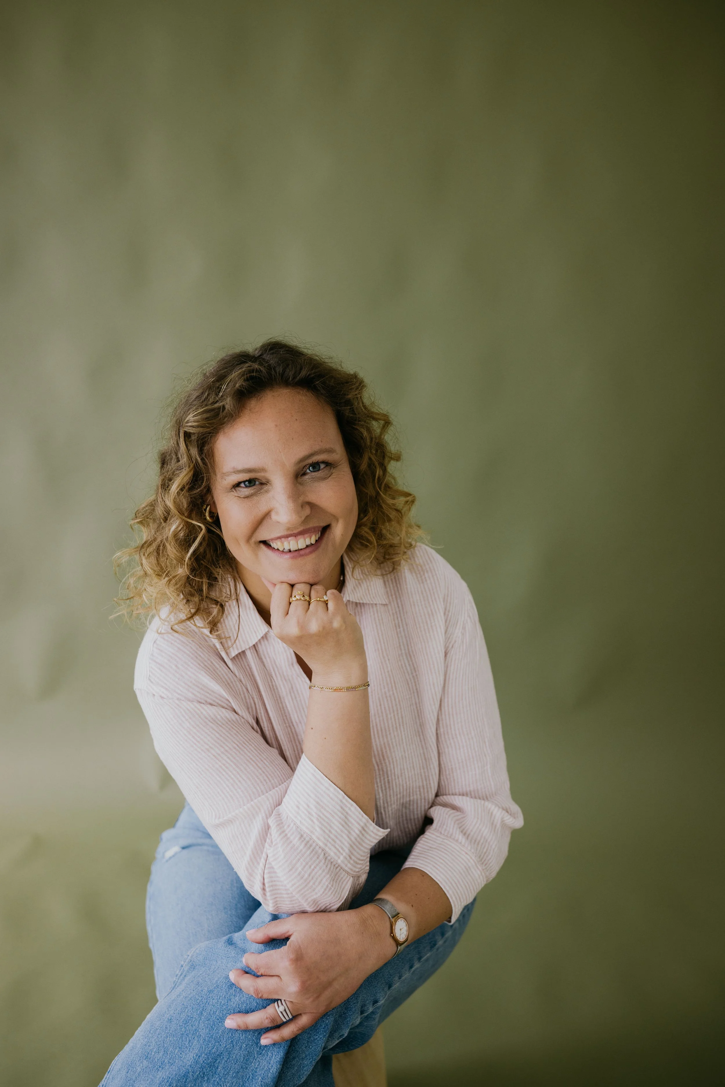 Een glimlachende vrouw met krullend haar in een lichte gestreepte blouse en blauwe jeans, kleermakerachtige houding met haar hoofd op haar hand, voor een groene achtergrond.