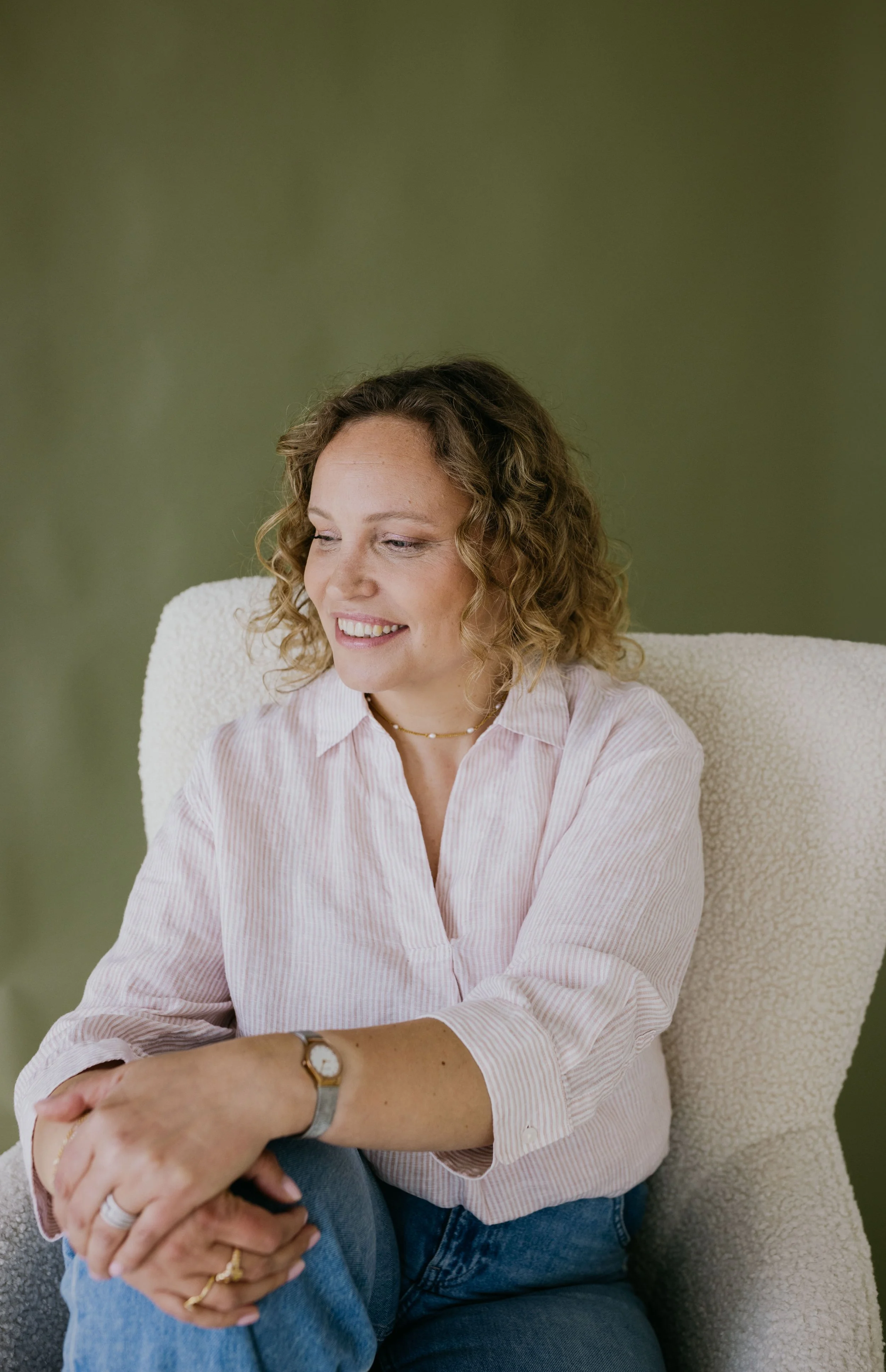 Een vrouw met gekrulde, schouderlange haar in een lichtroze gestreepte blouse zit in een stoffenstoel met een groene muur achter haar. Ze glimlacht en kijkt naar haar links, haar handen rusten op haar knie.