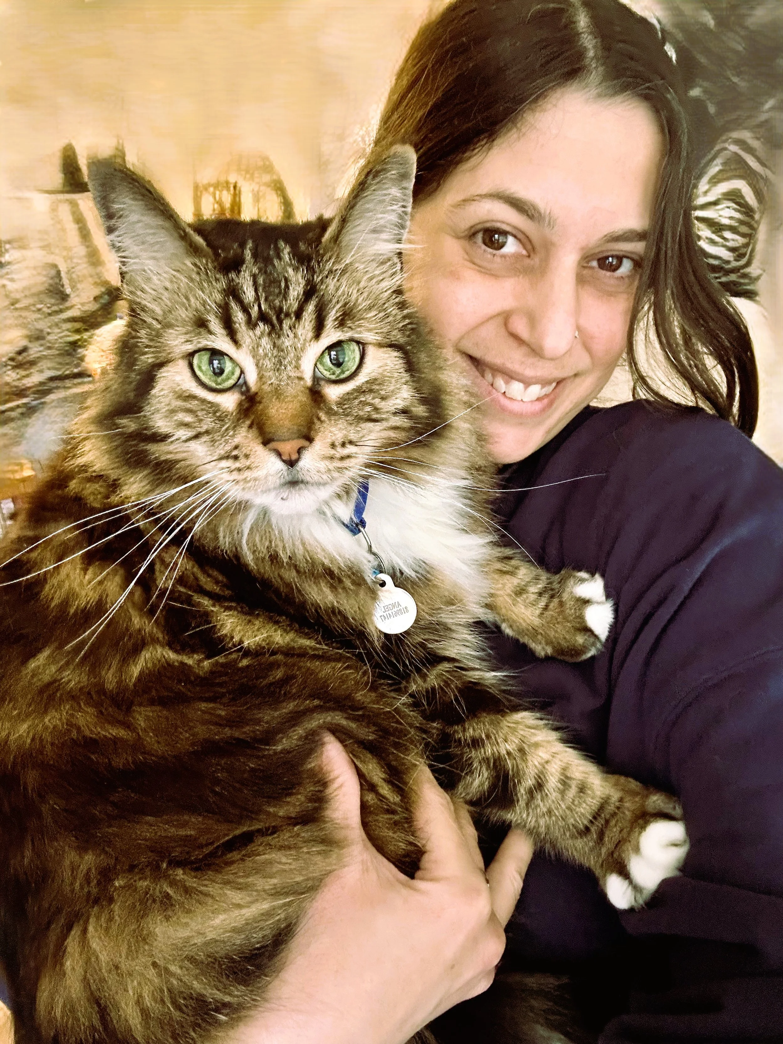 A woman with dark hair and a nose ring smiling while holding a large, fluffy tabby cat with green eyes close to her face.