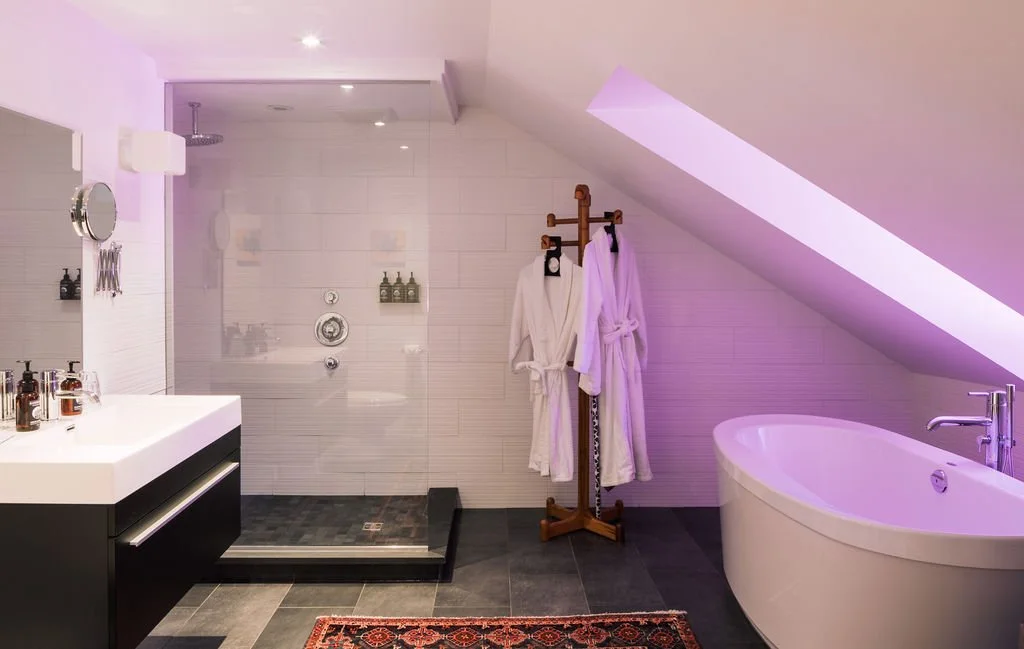 Modern bathroom with a walk-in shower, a white bathtub, a vanity with a mirror, and two white bathrobes hanging on a wooden coat rack. The bathroom has a sloped ceiling with a skylight and purple lighting.