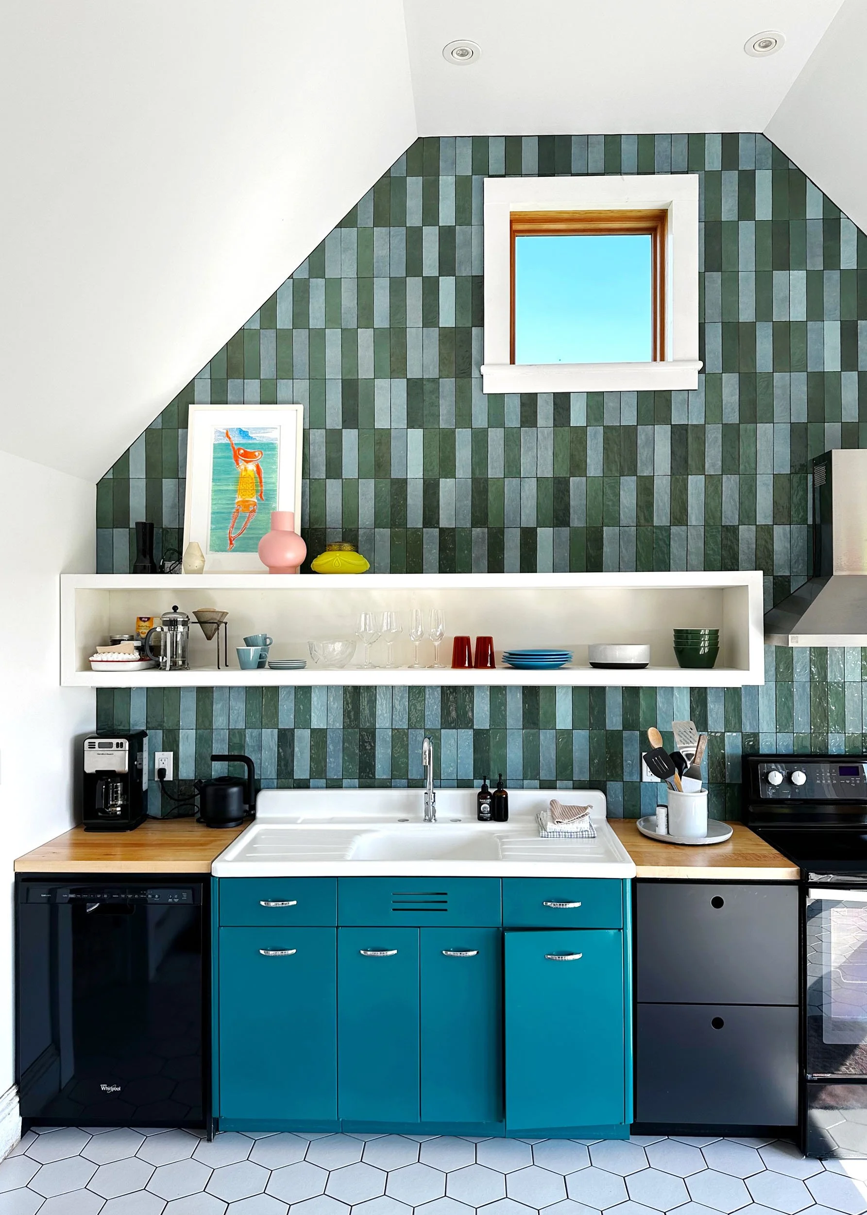 Kitchen with green tiled accent wall, blue lower cabinets, white shelves with dishes, coffee maker, and black appliances, chrome faucet, and a window with wooden trim.