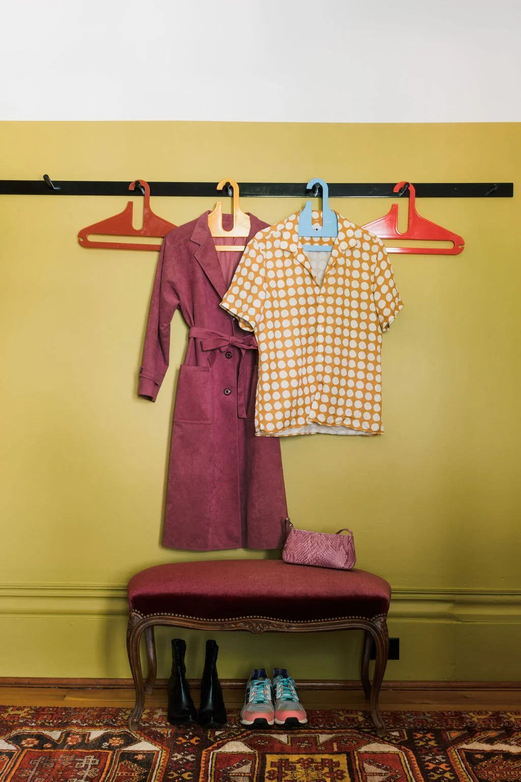 A pink coat, a yellow and white polka dot shirt, a pink clutch, black boots, and colorful sneakers are displayed against a yellow wall with a bench and a decorative rug.