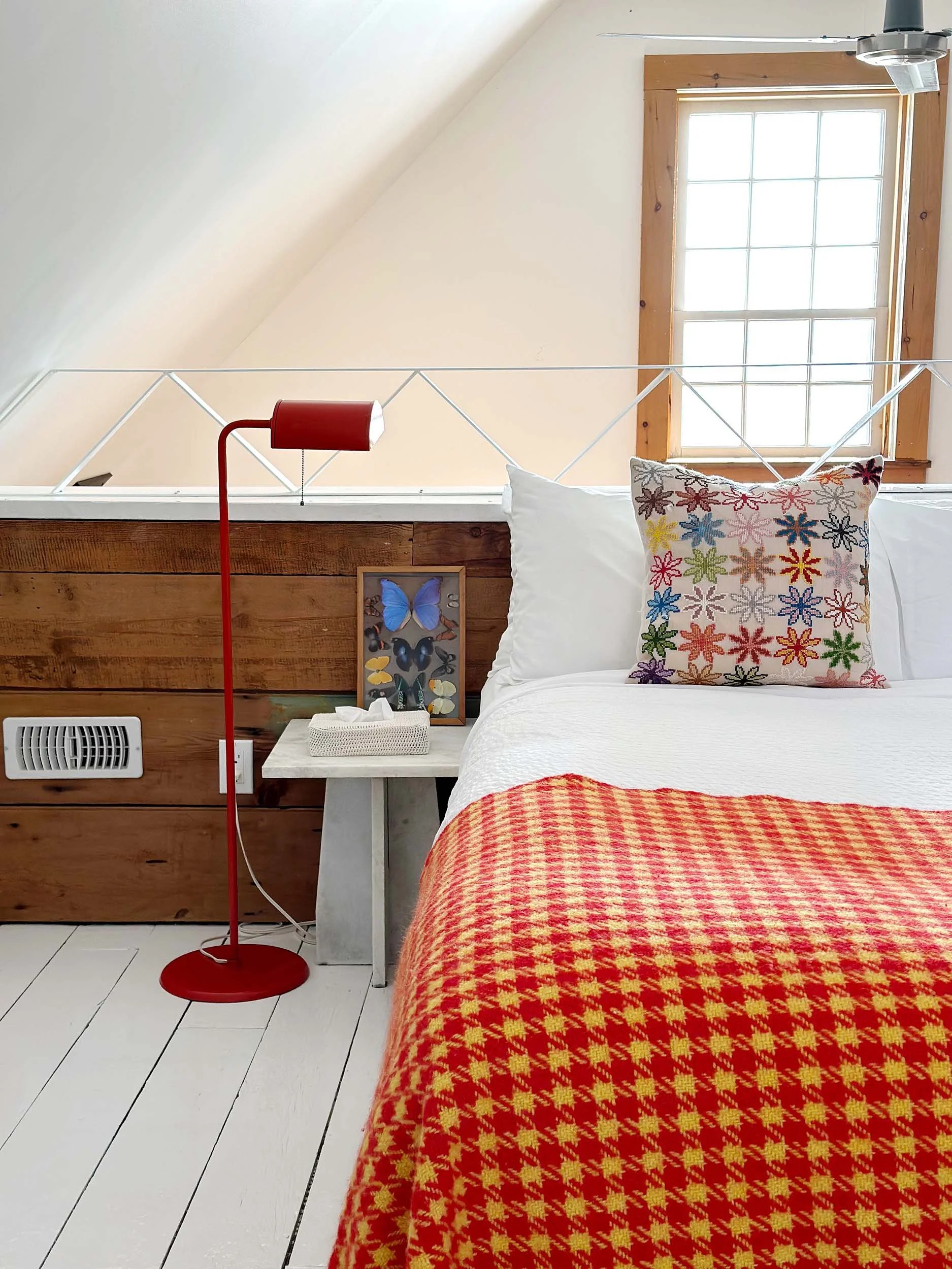 A bedroom with a large window, a red floor lamp, a white bedside table with a tissue box, a framed butterfly picture, pillows with embroidered floral patterns, a bed with a red and yellow patterned blanket, and a white wooden floor.