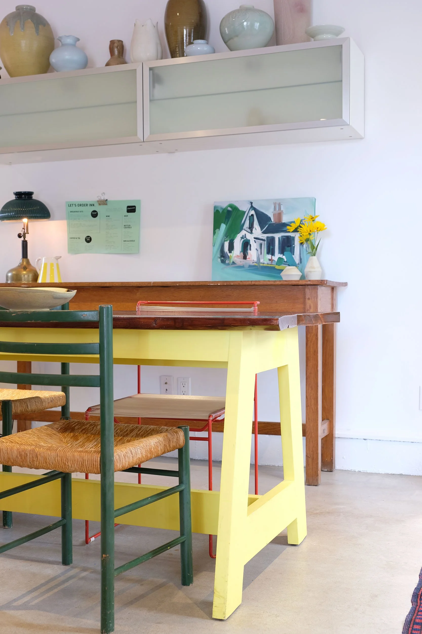 Colorful dining area with a yellow table, green and natural wood chairs, a painting of a house, and decorative vases and flowers on shelves.