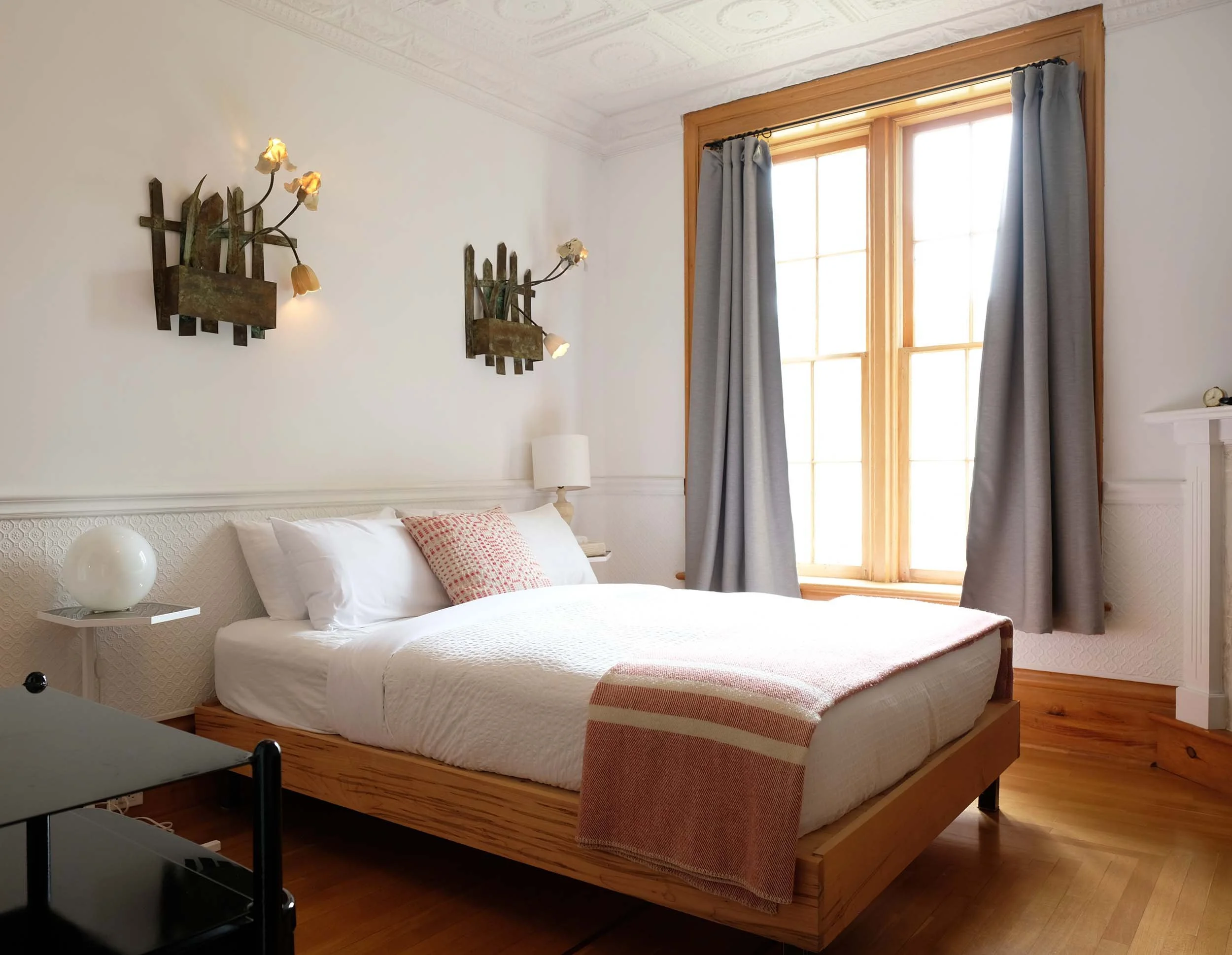 A bedroom with a large window with light blue curtains, a wooden bed frame with a white bedspread and a red and white patterned pillow, a white bedside table with a white orb lamp, wall-mounted light fixtures with flower-shaped bulbs, a clock on a white chest of drawers, and wooden floors.
