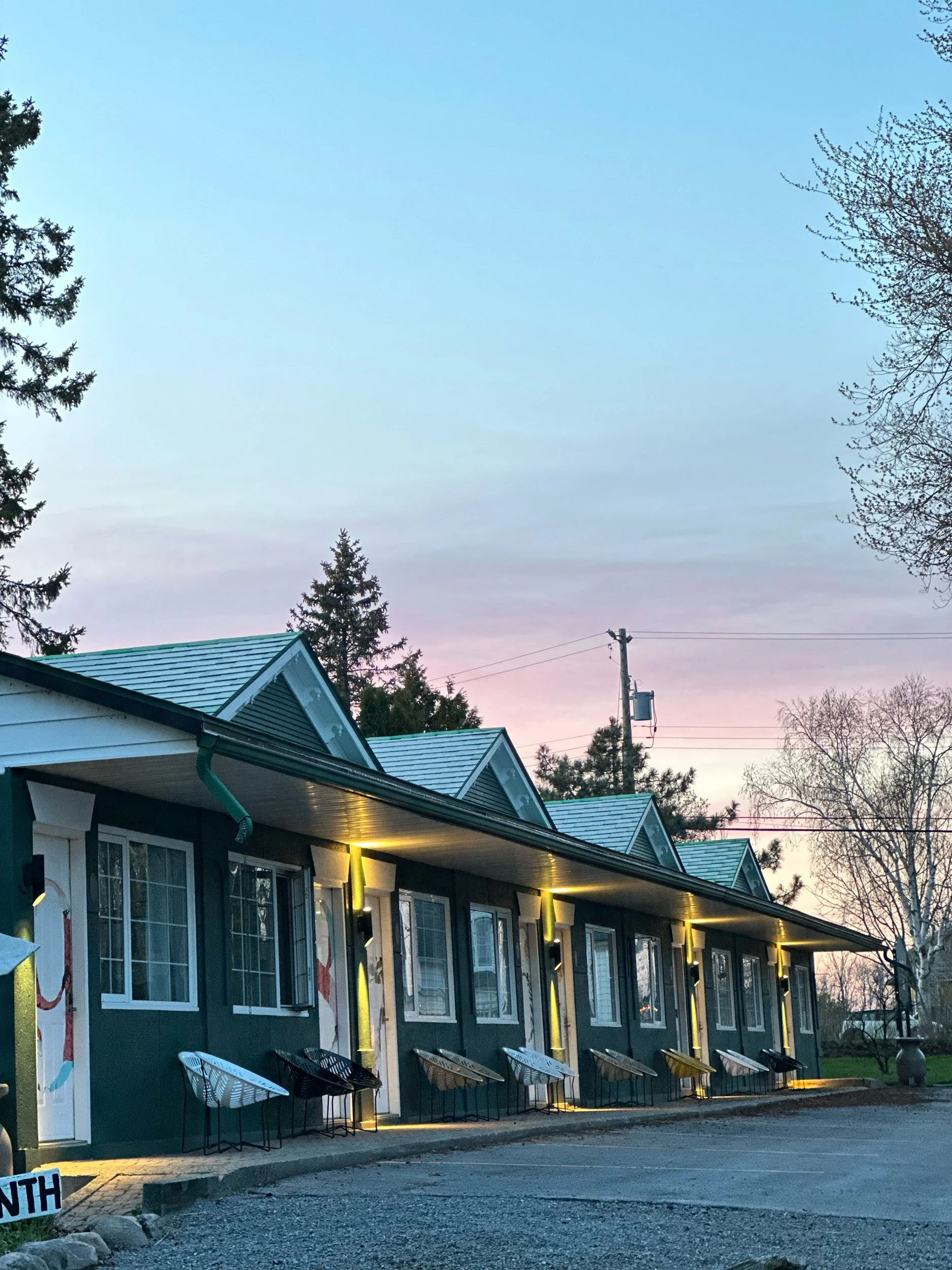 Exterior view of a row of apartment or hotel windows with yellow lights at dusk, neighboring the parking lot with a few chairs outside. The sky is cloudy with trees in the background.