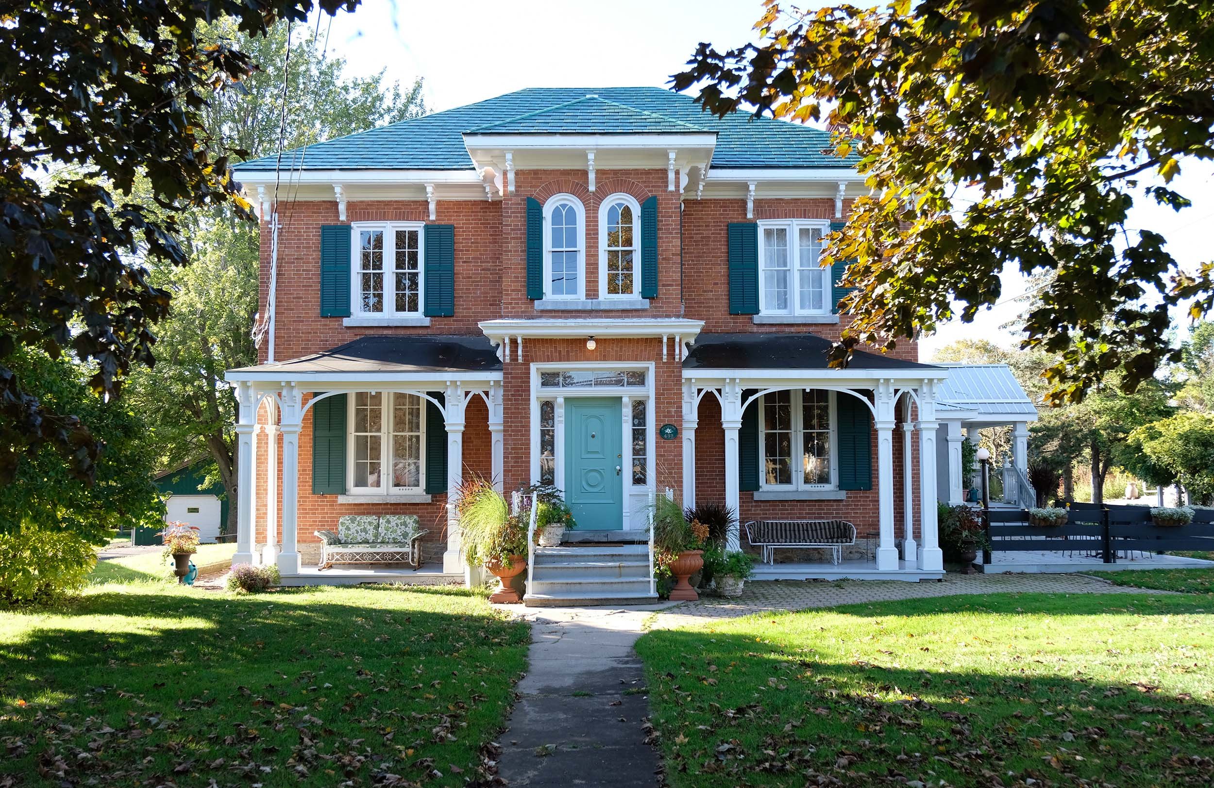 A large brick house with teal shutters and a teal door, surrounded by green trees and a lawn. There is a porch with white decorative trim, outdoor furniture, and potted plants.