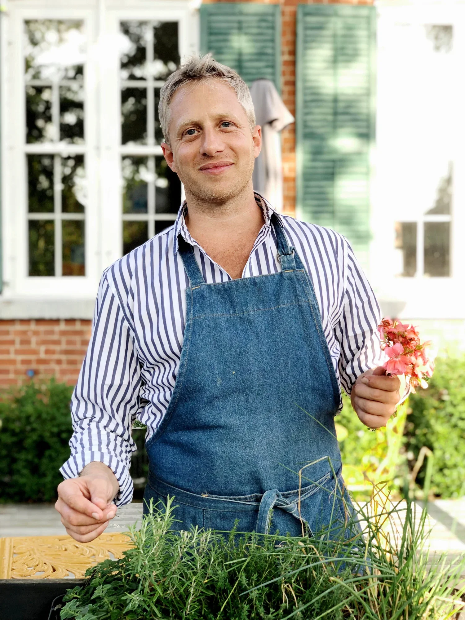 A man with gray hair wearing a striped shirt and denim apron is gardening outside, holding pink flowers while standing behind a table of various green plants.