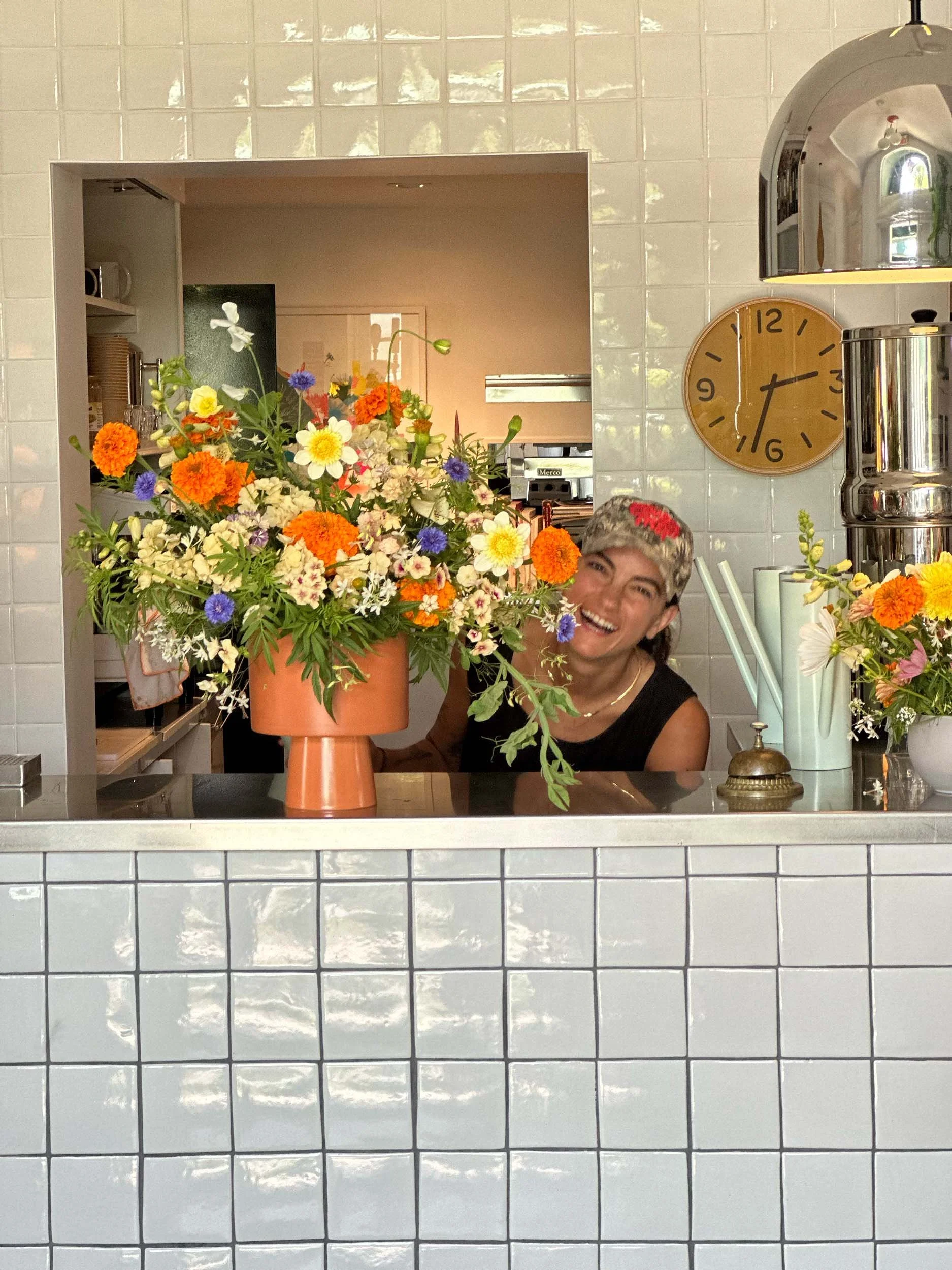 A woman with short hair and a big smile, wearing a black sleeveless top, is behind a counter in a kitchen. The counter has a large orange vase filled with colorful flowers including orange, white, purple, and yellow blooms. The background shows white tiled walls, a circular clock showing 1:13, and kitchen equipment.