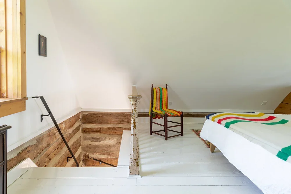 A cozy loft bedroom with white wooden floors, a bed with a colorful blanket, a wooden chair with a rainbow-striped blanket, and a staircase leading down to the lower level.