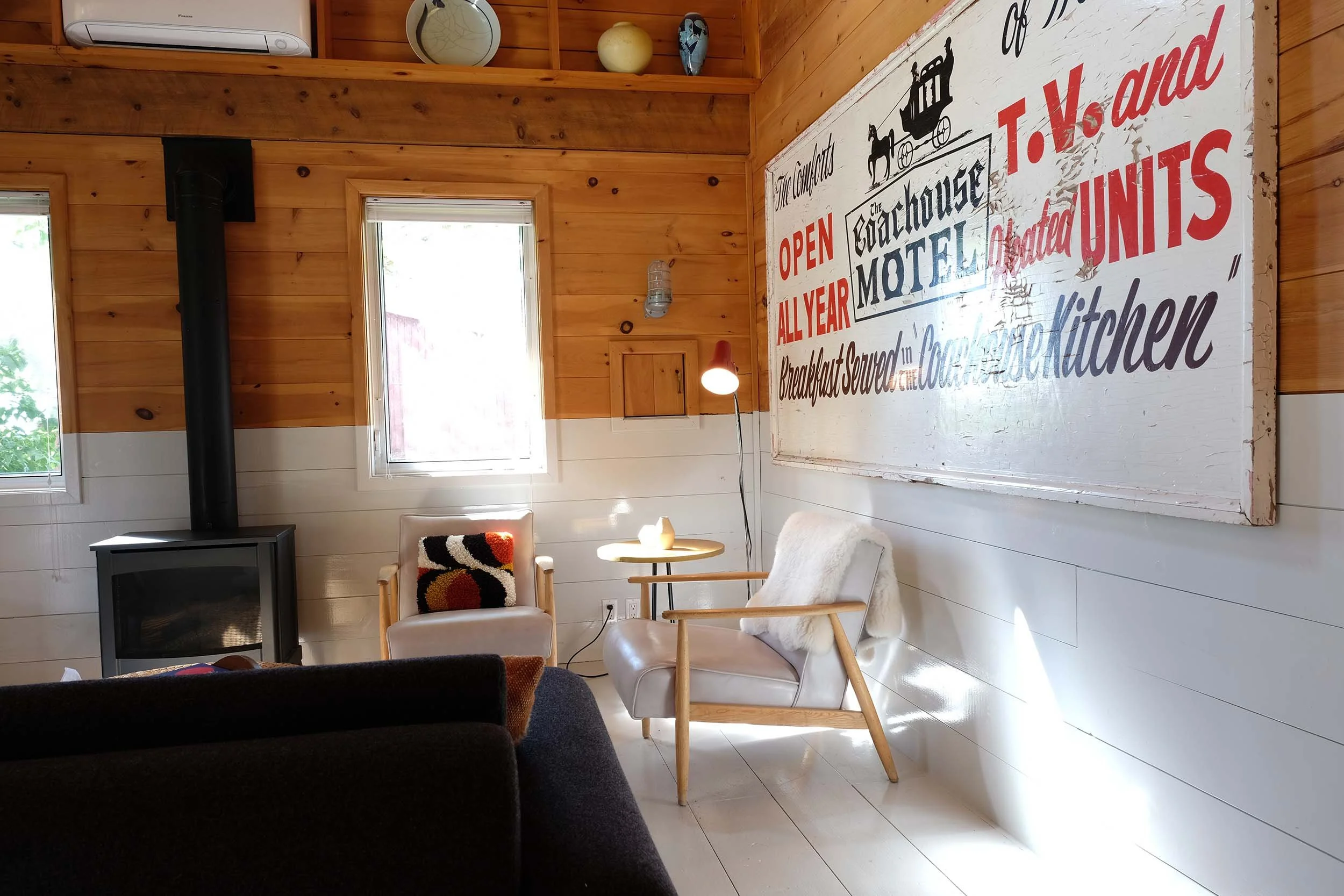 Interior of a cozy wooden room featuring a large vintage sign on the wall advertising a country inn and restaurant. The room has windows with blinds, a wood-burning stove, and a small sitting area with modern chairs and a lamp.