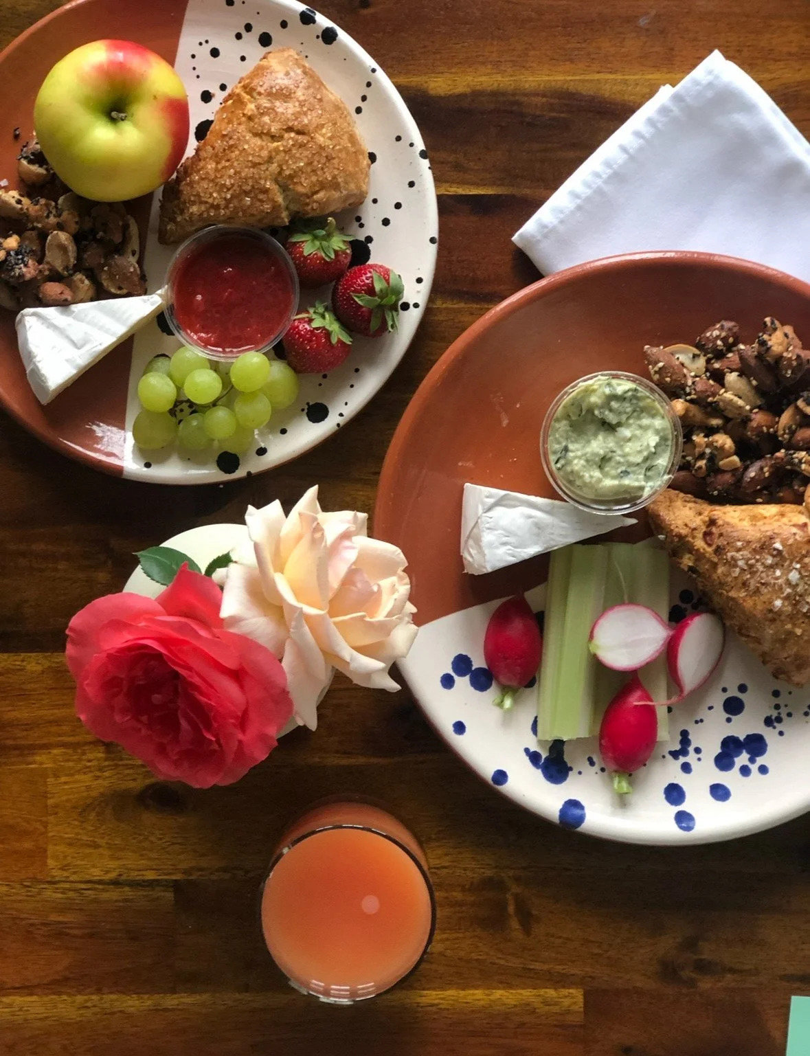 Two plates with fried food, mixed vegetables, and berries, along with small bowls of sauces, and a glass of pink juice, on a wooden table with flowers.