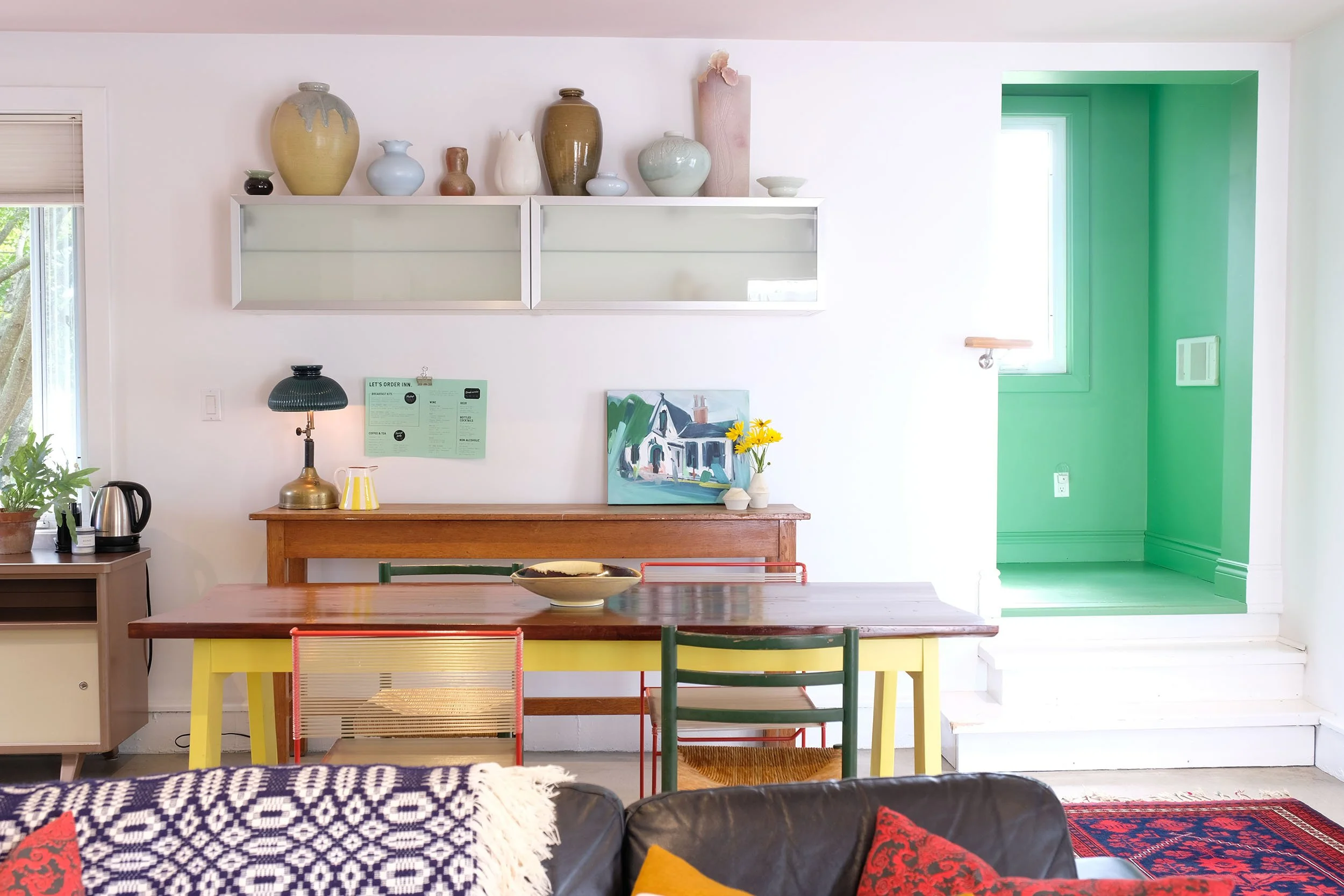 A dining area with a wooden table, colorful chairs, and a black sofa with red and yellow cushions. The wall behind has a large white shelf with decorative vases and a pink ceramic piece. A small table on the left holds a green lamp, a yellow vase, and a plant. There's a window with blinds and a painting of a house with yellow flowers on the right. A green doorway leads to another room, and part of a red patterned rug is visible at the bottom.