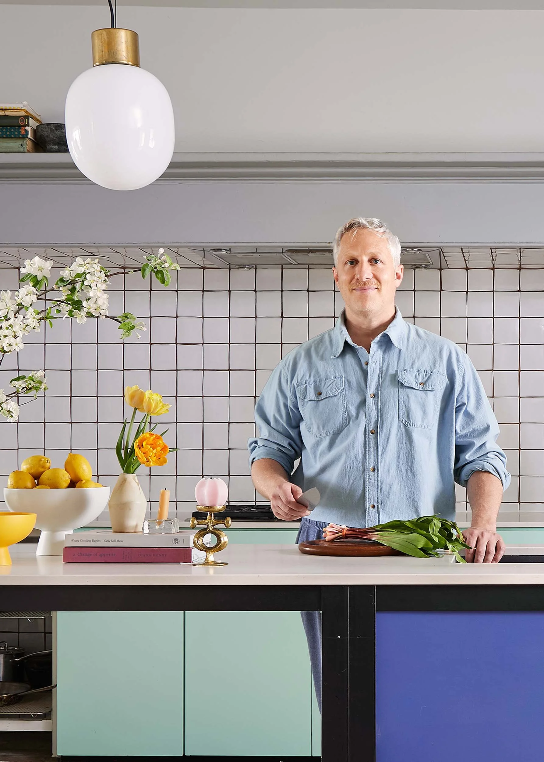 A man standing in a modern kitchen with a cutting board, green onions, a bowl of lemons, and flowers on the countertop. There is a white tiled backsplash and colorful cabinets below the counter. Overhead lighting with a large white and gold pendant lamp.