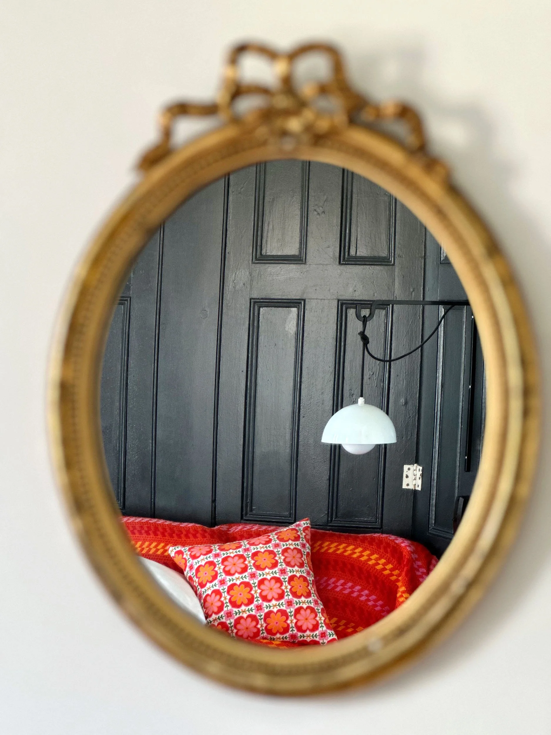 A mirror with a gold-colored decorative frame reflecting a black wooden-paneled wall, a hanging white globe lamp, and a red and orange patterned blanket or bedspread.