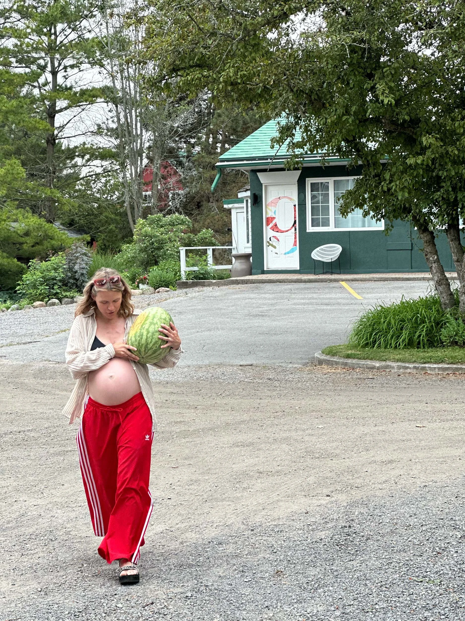 Pregnant woman walking outside holding a watermelon, wearing red pants and a beige shirt, with trees and a green house in the background.