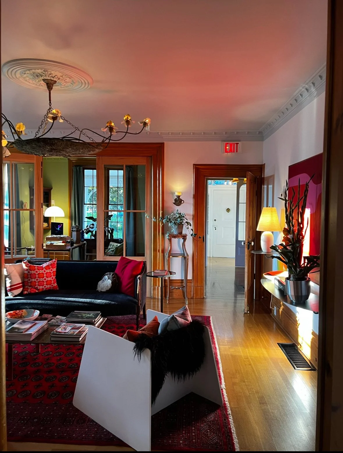 A cozy living room with a black sofa, red and patterned pillows, a wooden coffee table with magazines, and a white chair with a black furry pillow. There are warm lighting, plants, a chandelier, and a view into an adjoining room with green walls and large windows.
