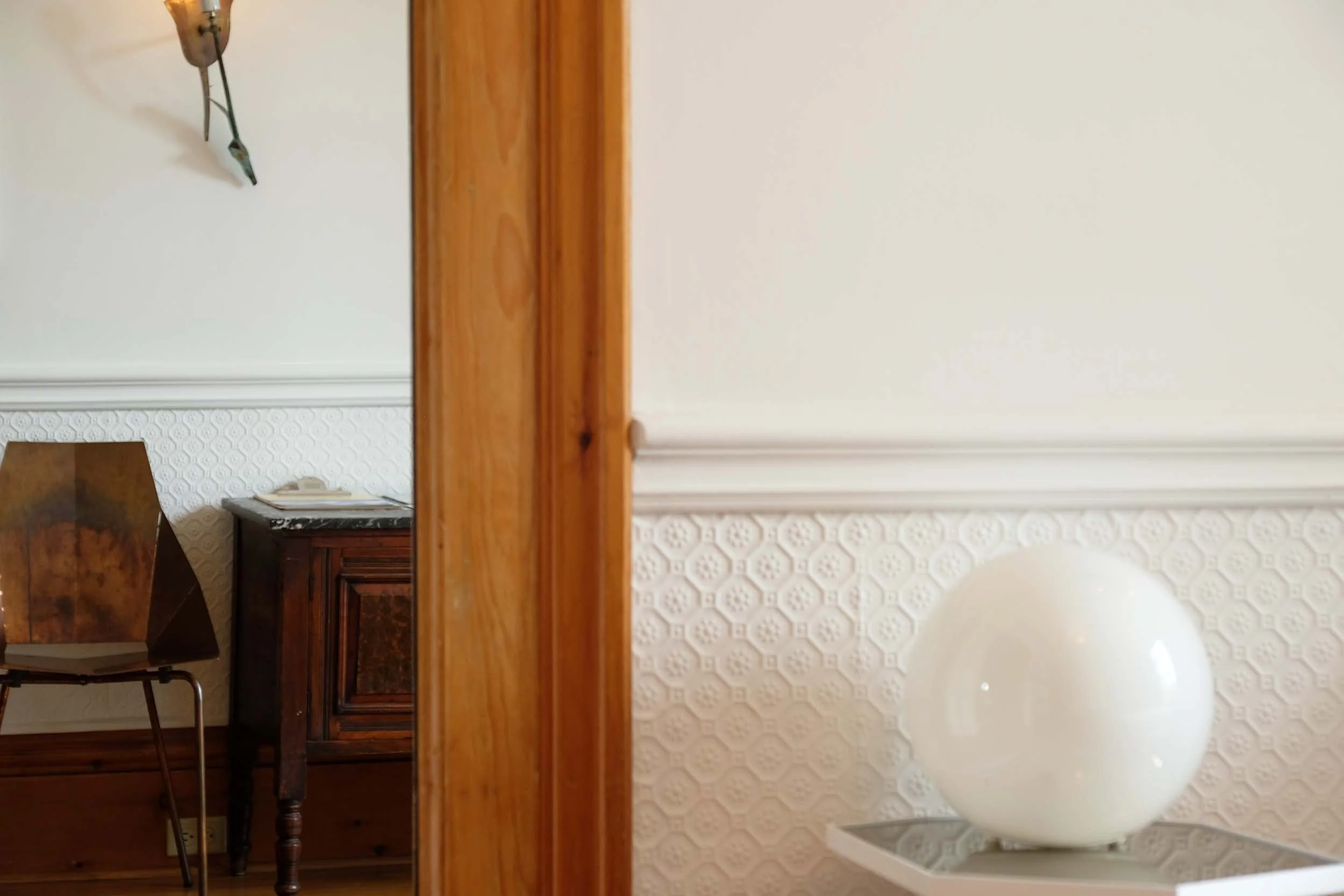 Part of a room with a wooden door or frame on the left. Inside the room, there is an antique wooden desk with a dark finish, a white wall with decorative molding, a textured white wallpaper with a geometric pattern, a chair with a wooden backrest, and a modern white spherical lamp with a narrow stand.