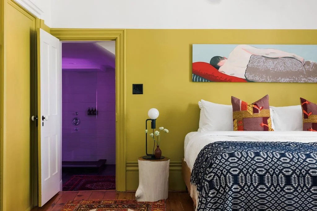 A bedroom with yellow walls and white ceiling, featuring a bed with white sheets, decorative pillows, and a black and white patterned blanket. Above the bed hangs a painting of a person lying on a pillow. To the left, there's a nightstand with a small lamp and a vase with white flowers. An open door reveals a purple-lit bathroom with a shower.