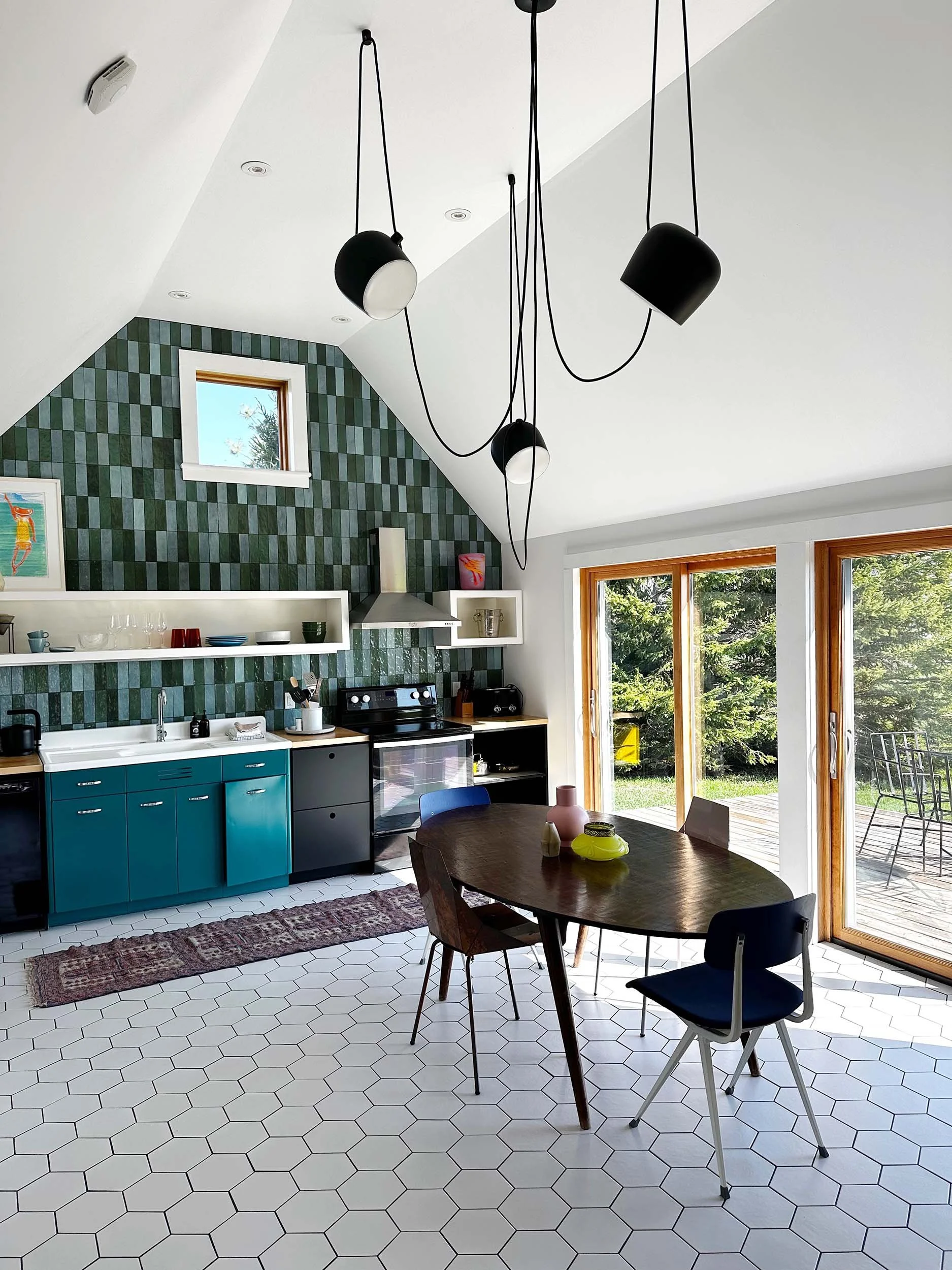 Modern kitchen and dining area with white hexagonal tile flooring, dark wooden dining table with chairs, teal lower cabinets, white upper and open shelves on a green tile accent wall, large windows revealing a green outdoor yard, and unique black pendant lights hanging from the ceiling.