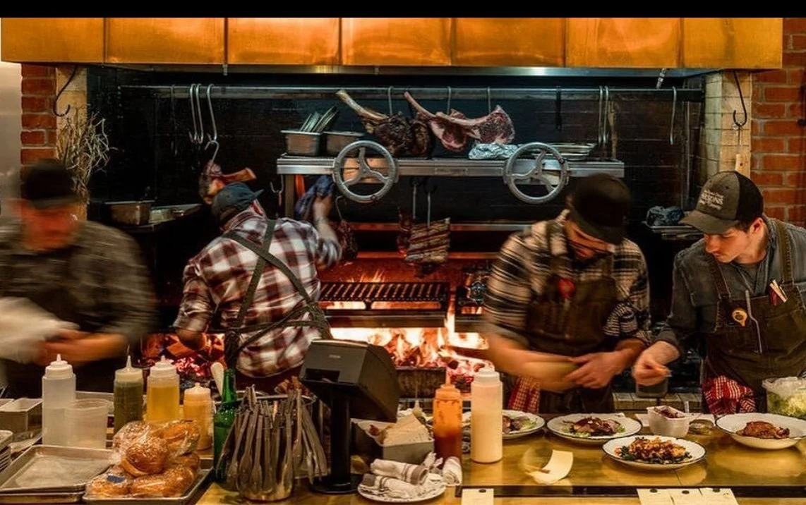 Four chefs working behind a restaurant counter with a brick fireplace, preparing food. The counter has various bottles, condiments, and dishes with prepared food.