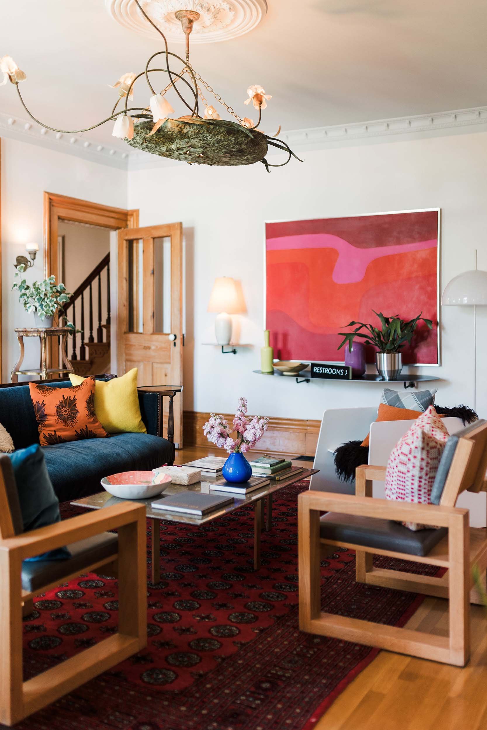 A living room with a red patterned rug, wooden chairs, a black sofa with colorful pillows, a coffee table with a blue vase and flowers, and wall art. The room has a chandelier and a wall sign indicating restrooms.