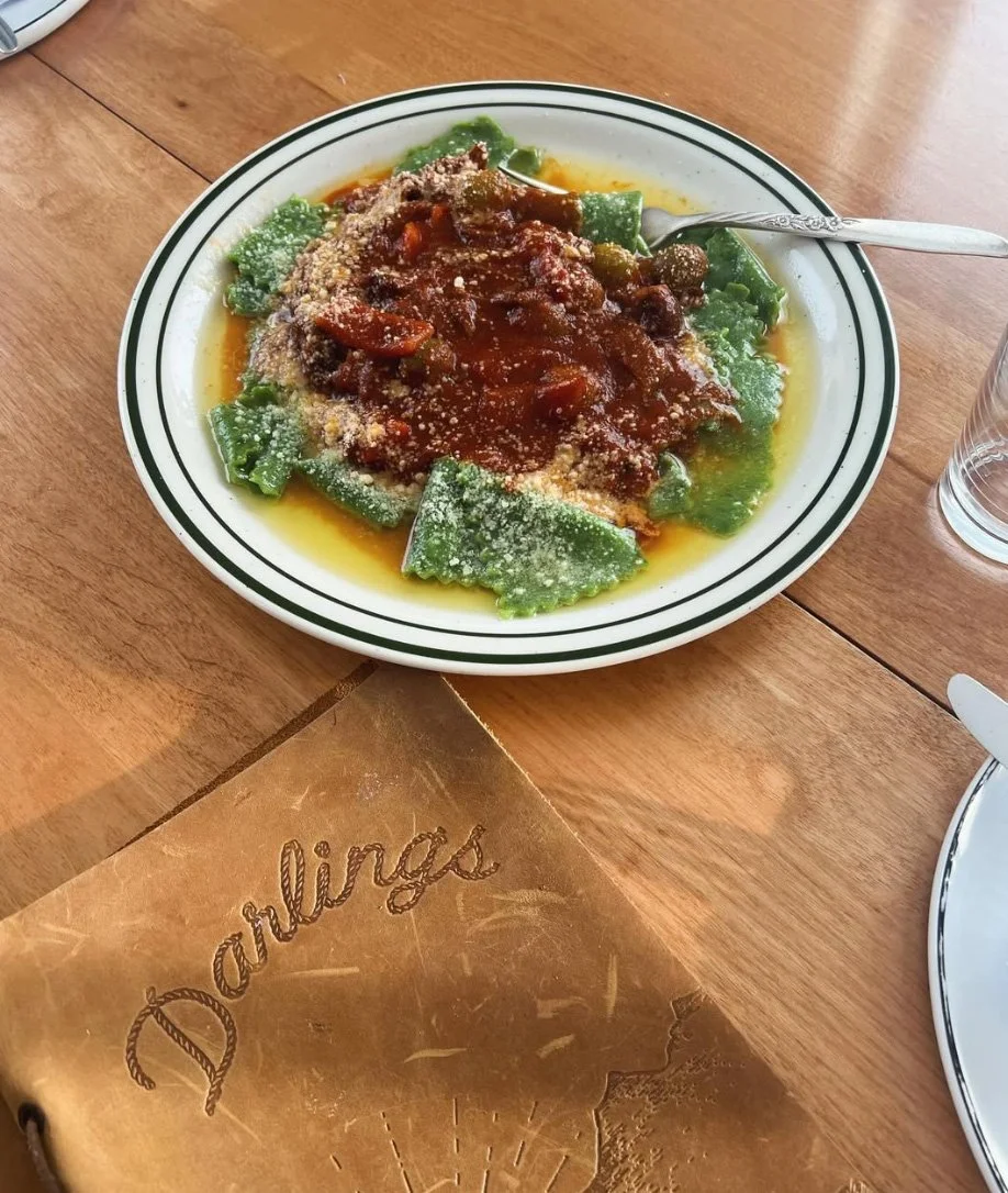 Plate of cooked ravioli pasta topped with tomato sauce, grated cheese, and green bell peppers on a wooden table, with a glass of water and a leather bill with the name 'Darlings' printed on it