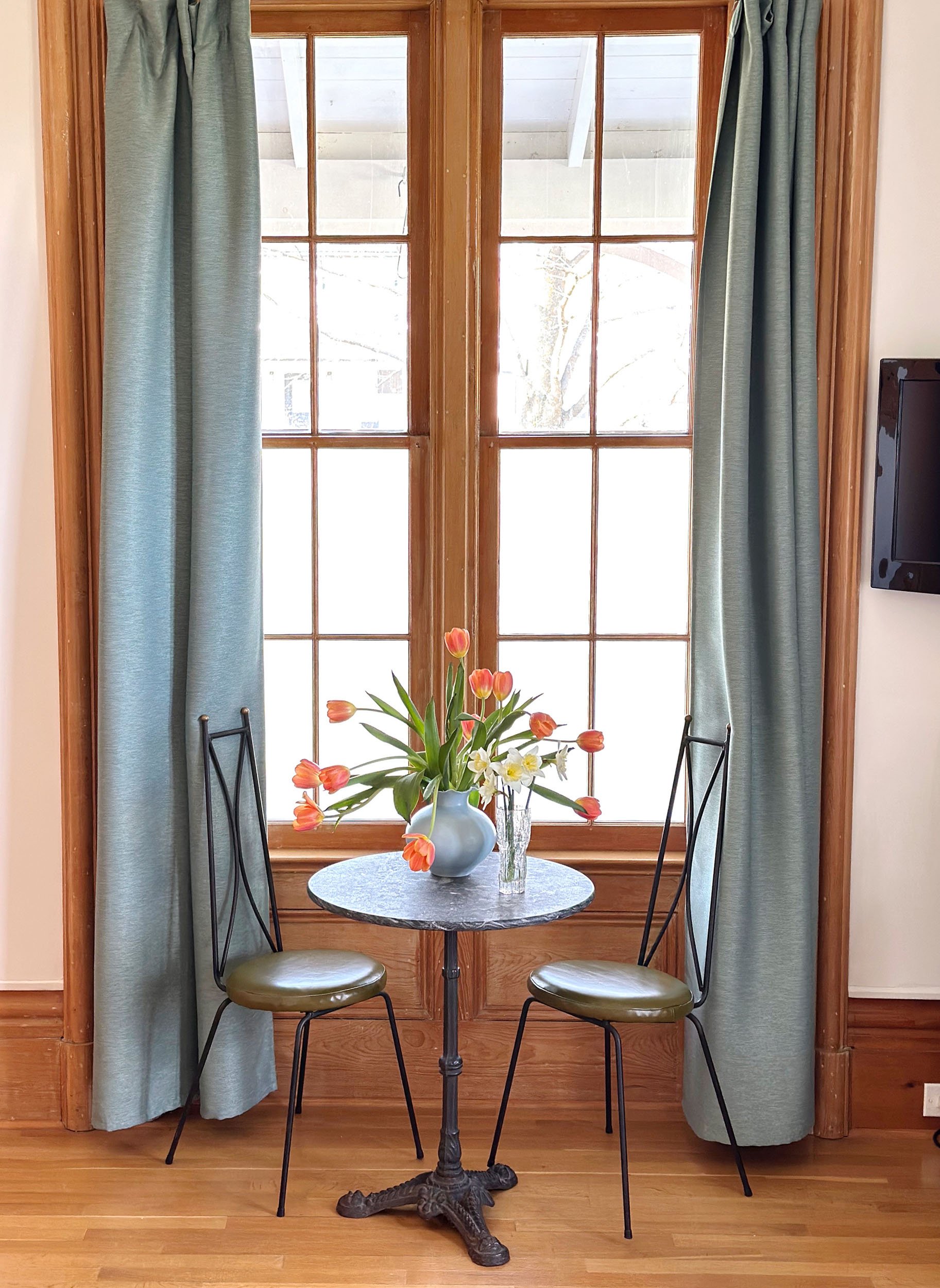 A small round table with a black ornate metal base, placed in front of a window with wooden frame and blue curtains. On the table, there is a white vase with orange tulips and a clear glass vase with white and yellow flowers. Two black metal chairs with green cushioned seats are positioned on either side of the table. The floor is wooden, and part of a TV screen is visible on the right side of the image.