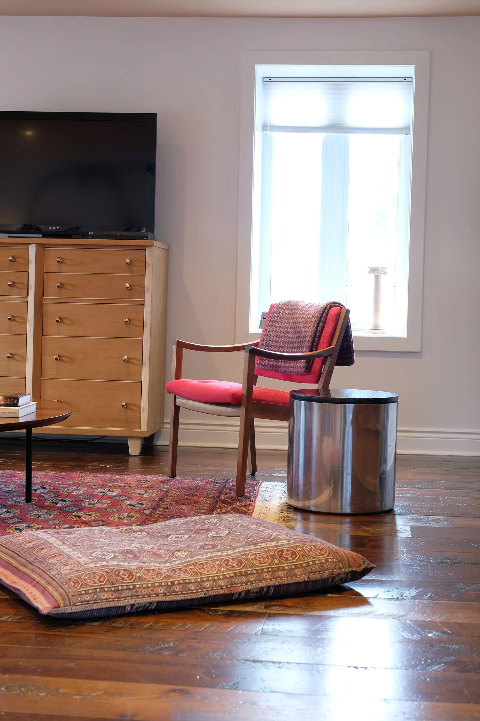 A cozy living room with hardwood floors, a patterned area rug, a wooden dresser with drawers and a TV on top, a chair with a red cushion and patterned back, a small round table, and a stainless steel trash can near the chair. There is a window letting in bright sunlight