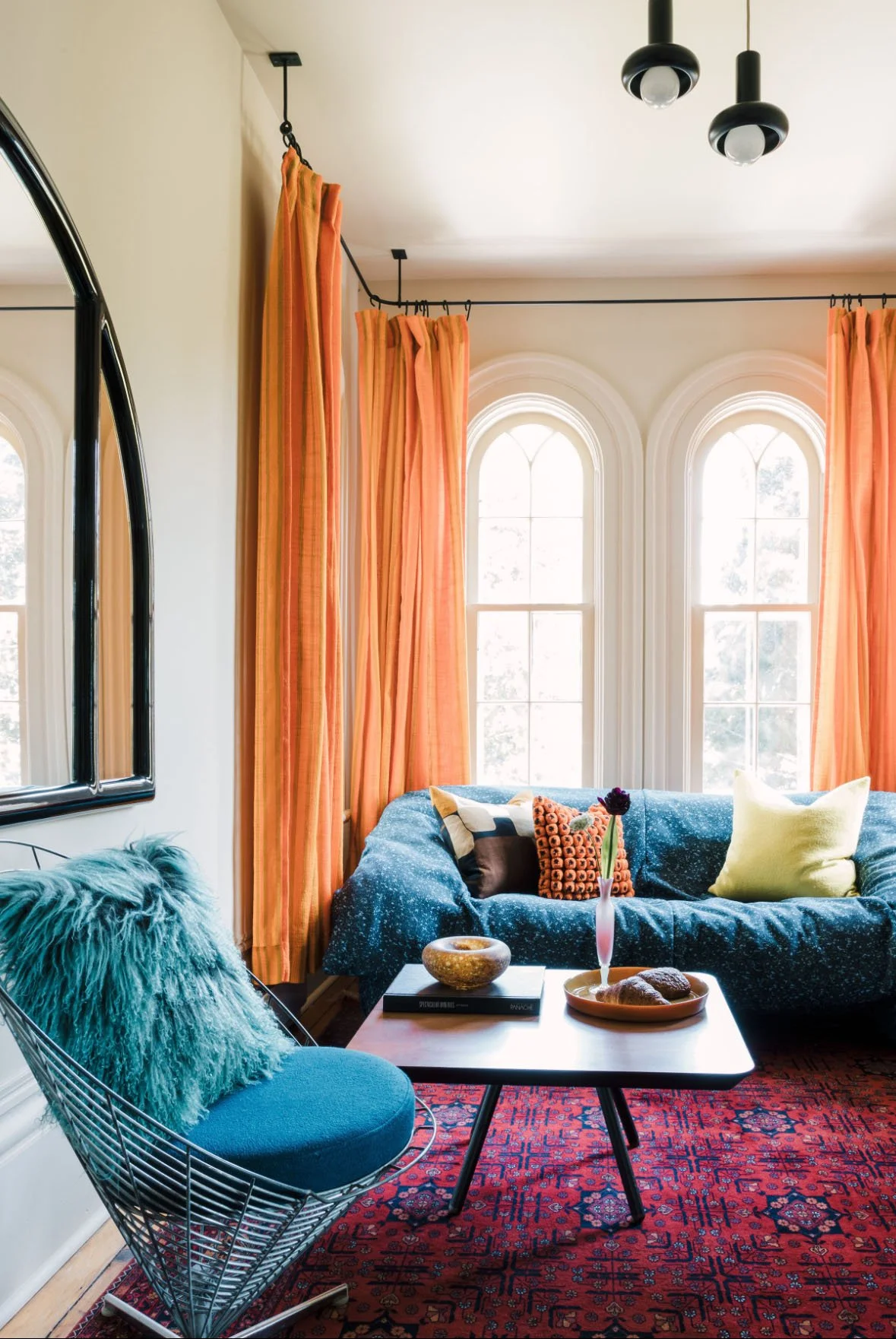 Living room with a blue sofa, colorful throw pillows, and large arched windows with orange curtains. There is a round coffee table with a vase of flowers, a bowl, and books. The room features a patterned red rug, a mirror on the wall, and a black wire chair with a teal fuzzy pillow.