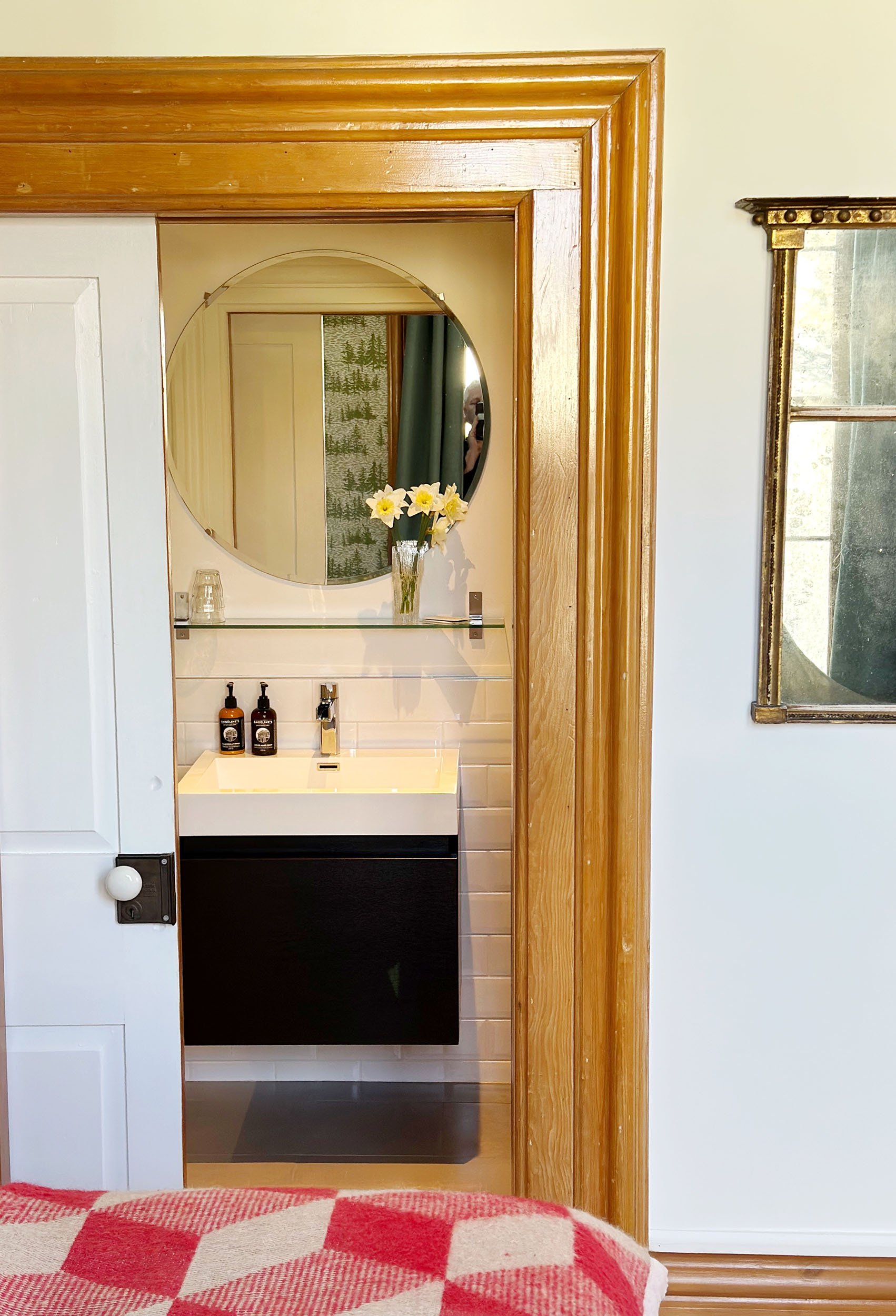 A small bathroom with a white sink, a round mirror, and a vase with white flowers. The bathroom is framed by a wooden door frame, with a window visible to the right.