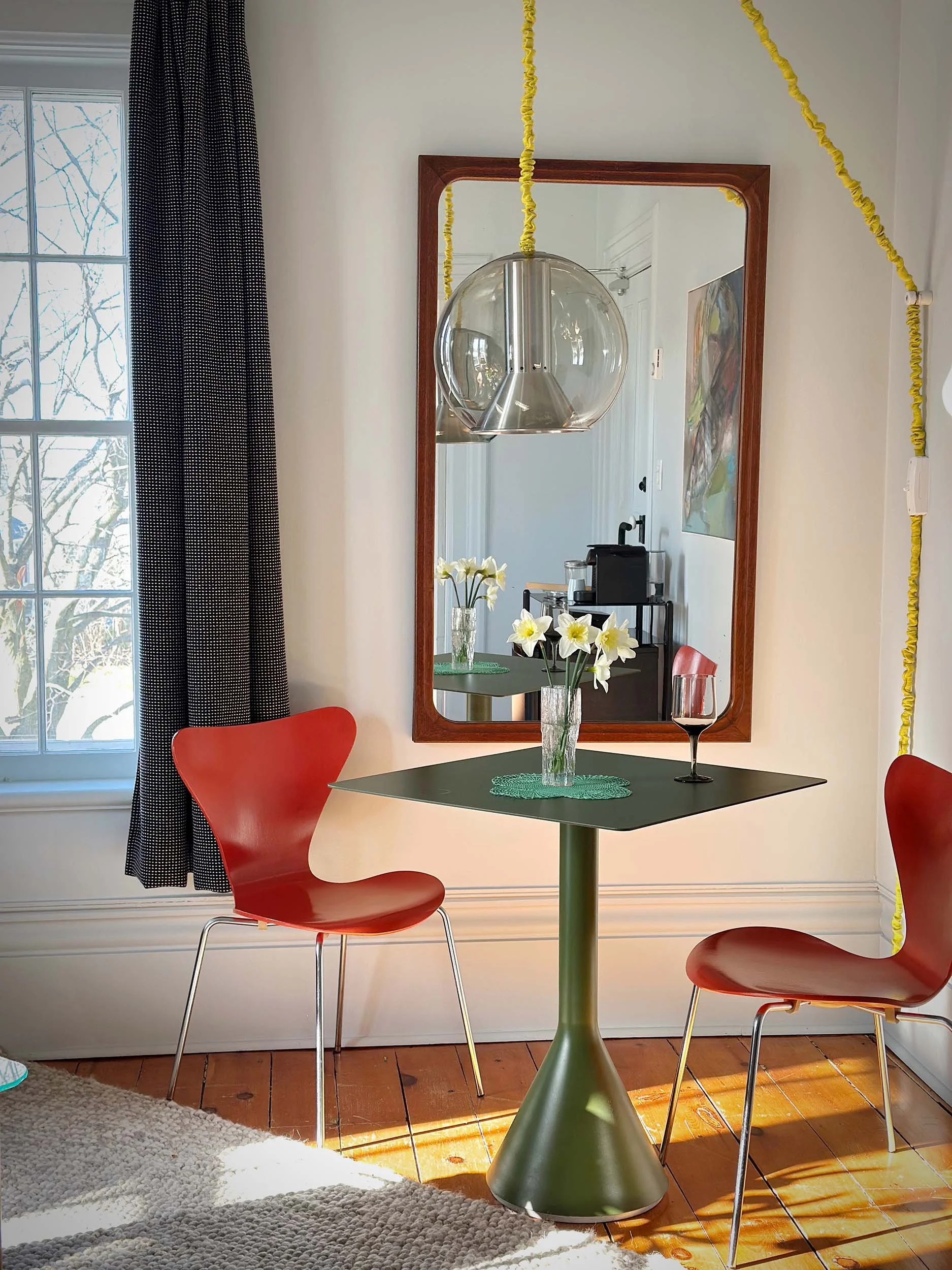 A dining room with a green table, three red chairs, a vase with white flowers, a glass of red wine, a mirror on the wall, a window with black and white curtains, a hanging light fixture, and a picture on the wall. Sunlight is streaming in, illuminating the wooden floor.