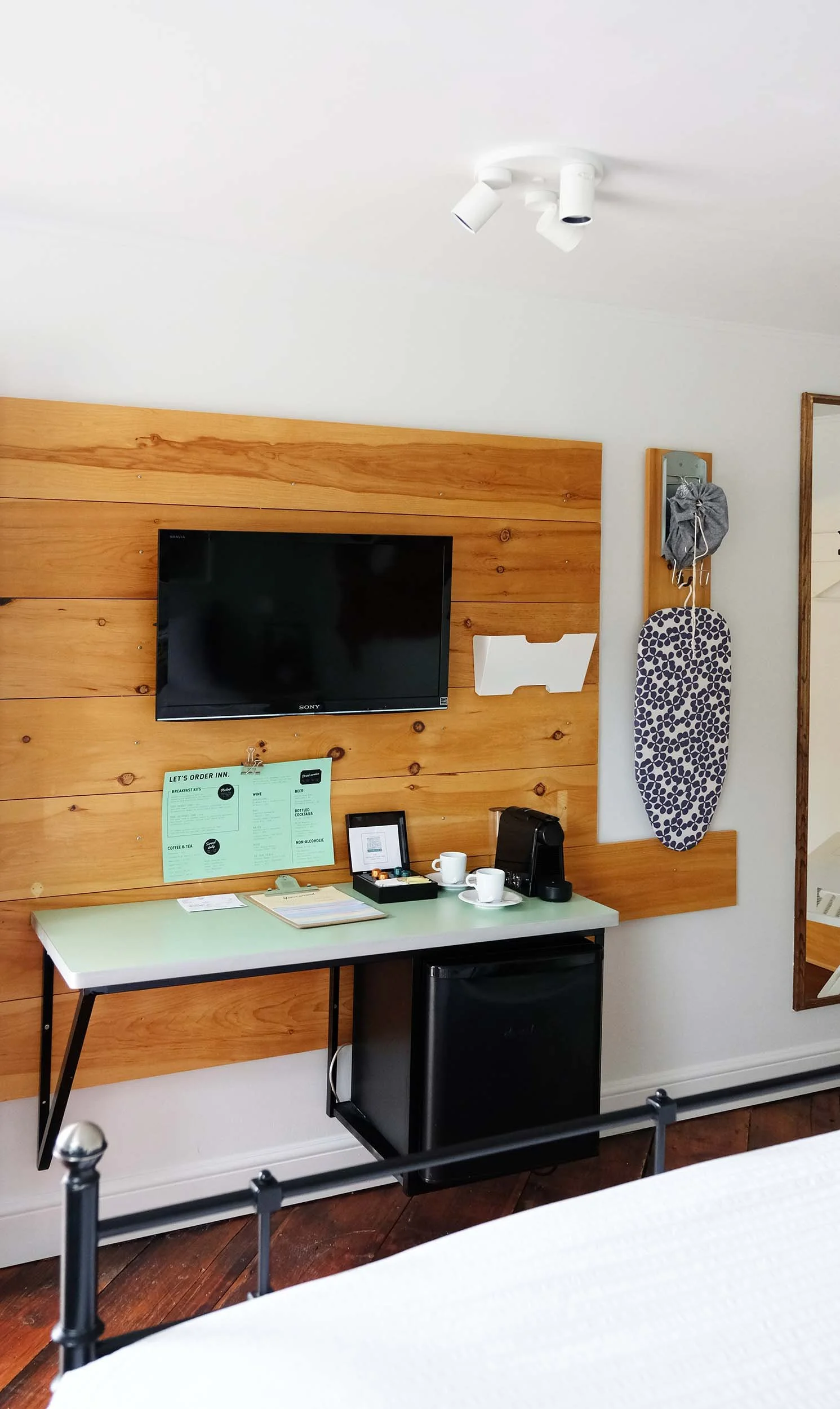 A hotel room with a mounted flat-screen TV on a wood-paneled wall. Below the TV is a table with a coffee maker, cups, and a small refrigerator. To the right, a mirror with hooks for hanging items, including a gray cloth bag and a patterned ironing board. The ceiling has a white light fixture with three adjustable spotlights.